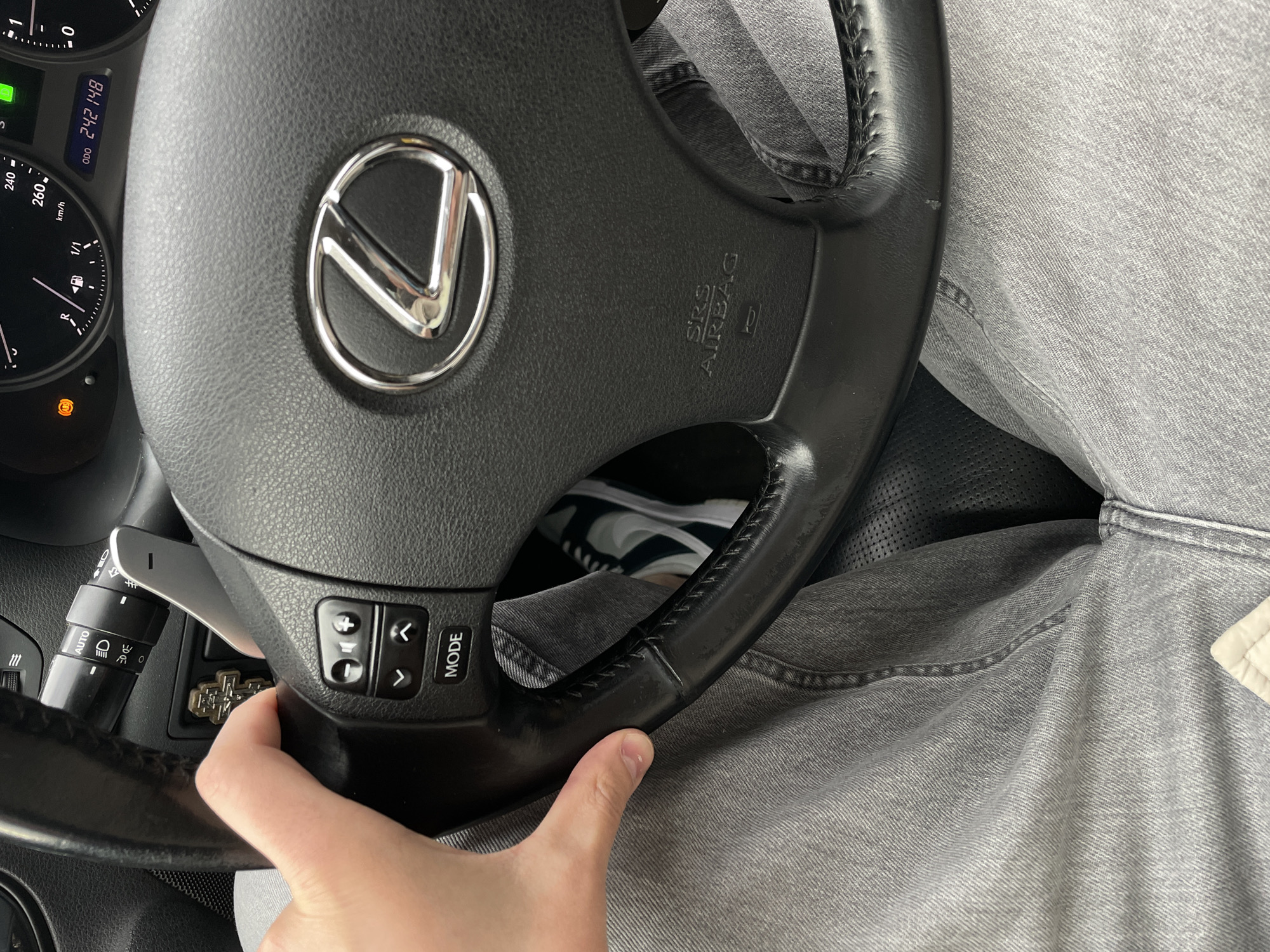 The image shows the interior of a Lexus vehicle. A hand is visible on the steering wheel, which features the Lexus logo and controls for audio and other functions. The dashboard includes a speedometer and an odometer reading 242148. The driver is wearing light gray jeans. Part of a sneaker is visible below the steering wheel. There is no information that would indicate the location is the city of Krasnoyarsk, Russia.