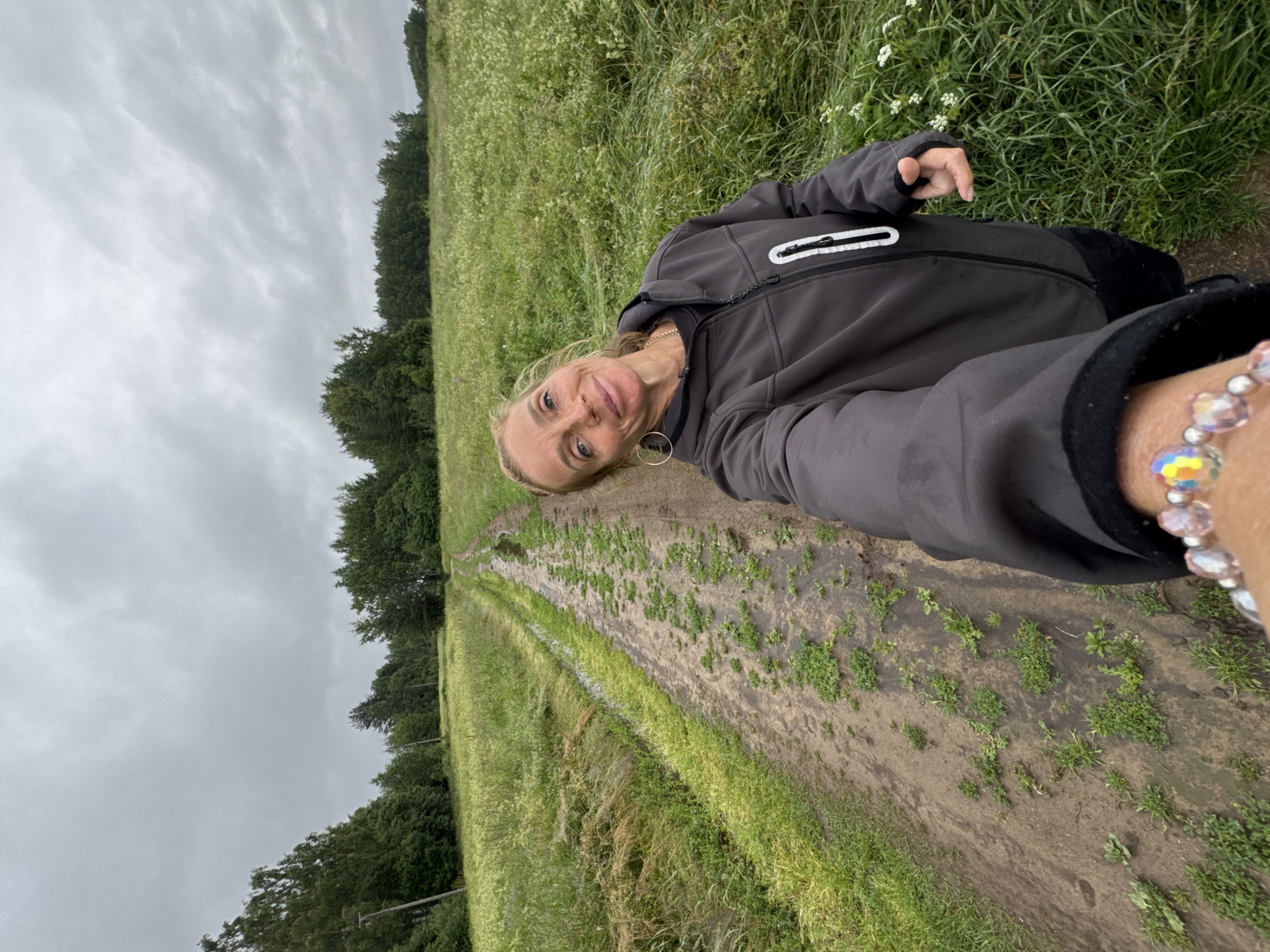 The image shows a woman taking a selfie outdoors. She is wearing a dark gray jacket with a white zipper and a black collar. A beaded bracelet with colorful beads is visible on her right wrist. She has blonde hair and is smiling at the camera. The setting appears to be a rural or natural environment with a path through a field of grass and some scattered plants. Trees are visible in the background on the left side of the image, and the sky is overcast. The path seems to be made of dirt and has some vegetation growing in it. The time of day is not definitively clear, but the overcast sky suggests it might be daytime, possibly late afternoon or a cloudy day. The location is suggested to be Mars, Russia, which is a notable detail in the context of the image. No visible text is present.