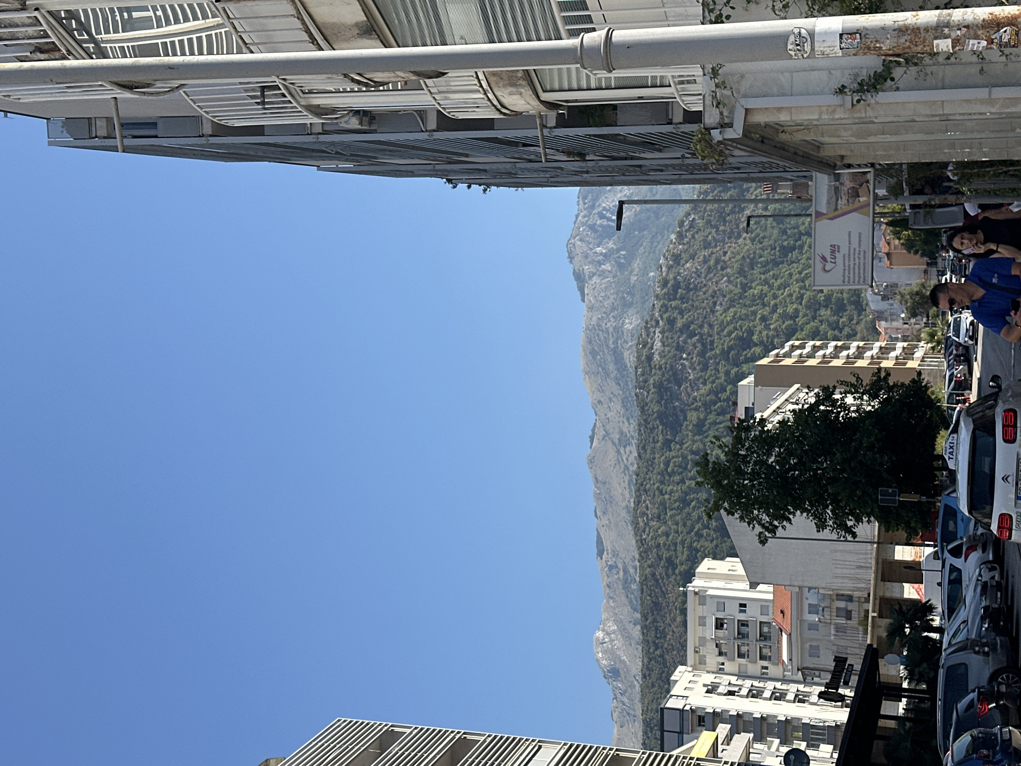 The image depicts a street scene in Budva, Montenegro, with a clear blue sky overhead. In the foreground, a street is lined with buildings, and parked cars are visible. To the right, a modern apartment building with balconies dominates the view. On the left, another apartment building with white horizontal accents is present. In the background, a lush green hillside covered in trees rises towards majestic, rocky mountains. A billboard for "LUNA" is positioned on the side of the street. Two individuals, a man in a blue shirt and a woman in a black top, are walking on the sidewalk. The man is looking at his phone, and the woman is engaged in a phone conversation. The overall impression is a sunlit urban environment with a natural landscape as a backdrop.