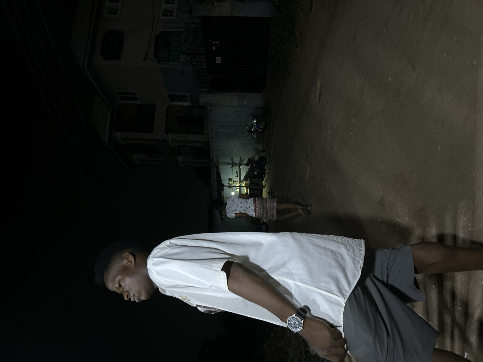 The image, apparently taken at night in Ihiagwa, Nigeria, depicts a man standing in the foreground, wearing a white short-sleeved shirt, dark shorts, and a watch on his left wrist. In the background, a woman is walking away from the camera, towards a building and a dimly lit commercial establishment. There are motorcycles parked by the building. The area appears to be a street or road. The lighting is poor, indicating minimal illumination from street lights or other sources. The building in the background is multi-storied. The road or street appears to be paved with asphalt.
