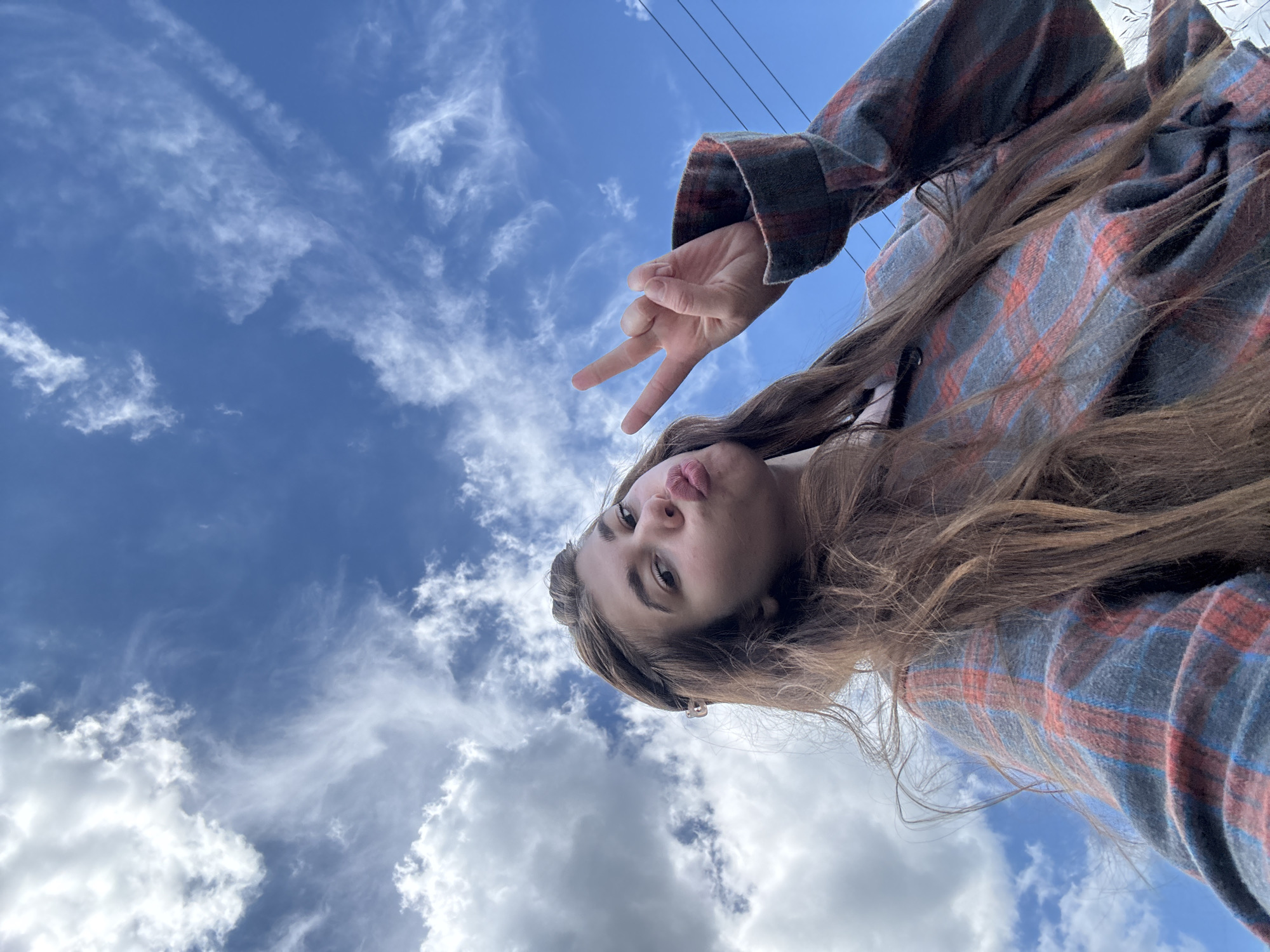 A young woman is outdoors, holding up two fingers in a peace sign. She is wearing a plaid shirt and has long brown hair. The sky behind her is blue with white clouds. The image does not provide any information about the location in the city of Мещёры, Russia.