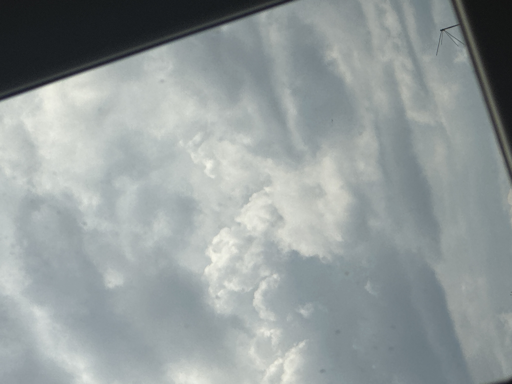 The image shows a view of the sky with various cloud formations. The image is framed by a dark, likely indoor, border suggesting the photograph was taken from inside a building or vehicle. An antenna-like structure is visible in the top right corner against the cloudy sky. The location is unknown.