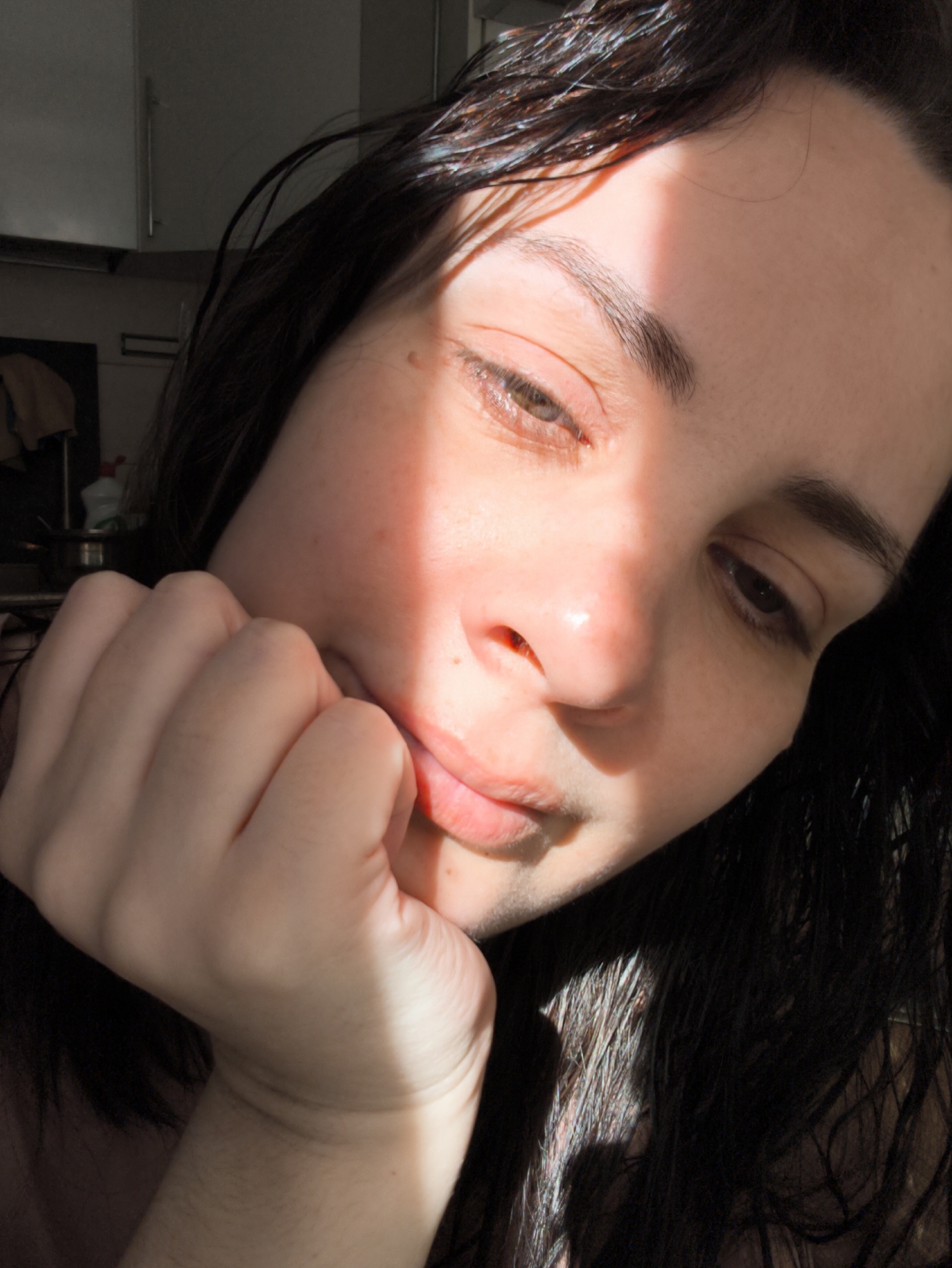 A close-up portrait depicts a person with dark, wet hair. The person is resting their chin on their hand, gazing downward. A beam of direct sunlight illuminates part of the face, creating a stark contrast between light and shadow. The background features a glimpse of a kitchen area with light-colored cabinets and cleaning supplies. The location of the image is in the city of Chaikovsky, Russia.
