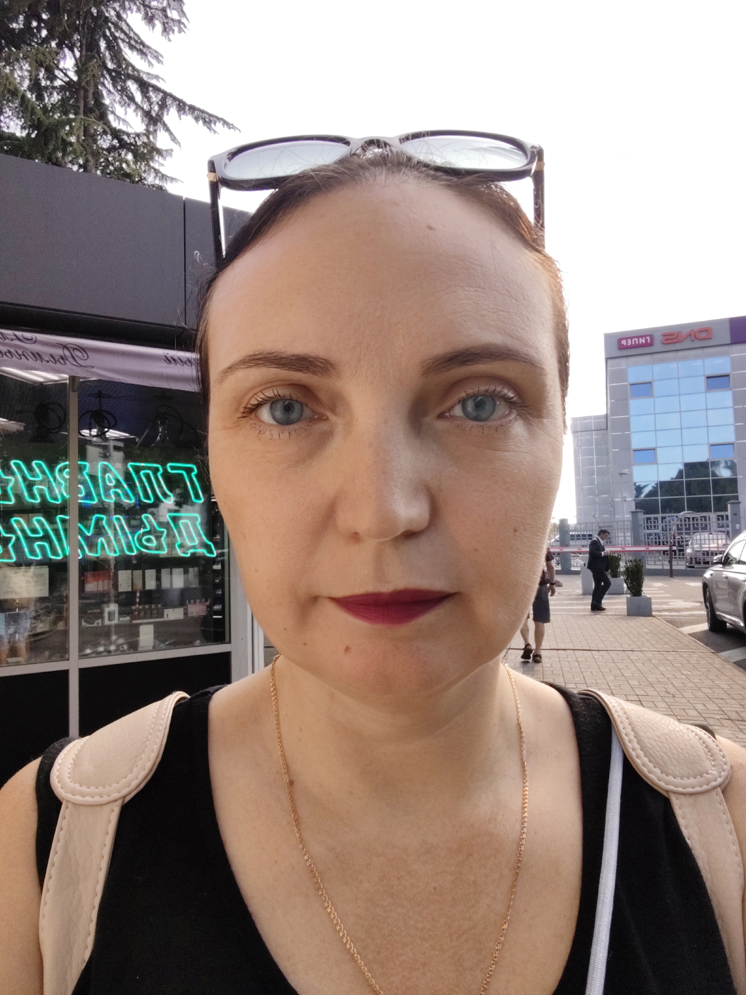 A medium shot photograph captures a woman standing outdoors, facing the camera. She has fair skin, blue eyes, and brown hair, with dark-rimmed glasses perched on top of her head. She is wearing a black sleeveless top with light-colored straps and a thin gold chain necklace. Her lips are painted a deep berry color.
In the background, to the left, a storefront features neon green lettering in a language that appears to be Cyrillic, possibly Russian. The storefront displays various products behind glass. Further to the right, a modern building with a glass facade and purple accents is visible. Several people are walking on a paved walkway in front of this building, and a silver car is parked on the right side of the frame. Trees are present in the upper left portion of the image. The lighting suggests it is daytime, likely in Sochi, Russia, given the architectural style and the Cyrillic signage.