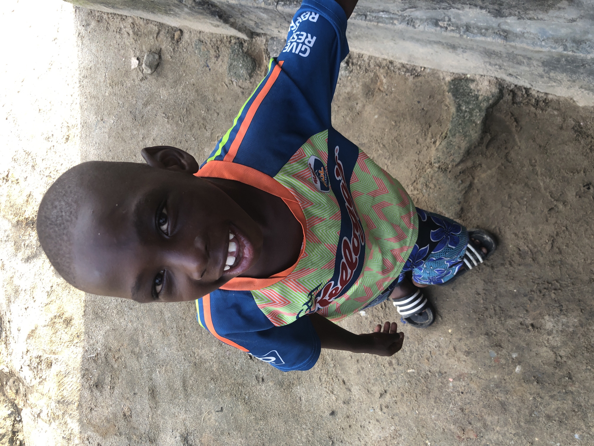 A high-angle medium shot captures a young Black boy smiling broadly while standing on a dirt ground in Ado, Nigeria. He is wearing a blue and green jersey with orange trim and a blue and floral pattern on the shorts. His black hair is shaved short. He is wearing black and white striped flip-flops. A concrete wall is to his right. The ground is a mix of sand and dirt with scattered small rocks and debris. The image is taken in daylight.