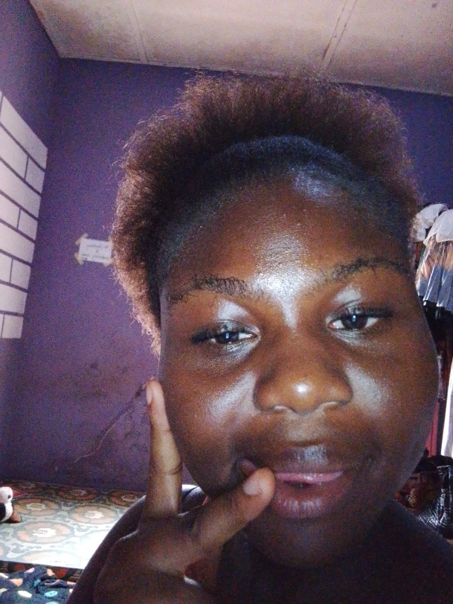 A close-up, selfie-style photograph depicts an adult female of dark skin complexion, centered in the frame. Her dark, textured hair is styled upwards, possibly secured by a band. Her eyes are open and directed at the camera, and her lips are slightly parted, revealing a portion of her tongue, indicative of a subtle smile. Her right index finger is bent and pressed against her lower lip, with her thumb also contacting the lip. Her skin surface exhibits a visible sheen. The background reveals an indoor room with solid purple walls. To the upper left, a section of white horizontal slats or a window blind is visible. A small, light-colored rectangular object is affixed to the purple wall behind the subject's left side. The floor in the lower-left corner features a patterned surface, appearing to be a mat or linoleum with a repeating light-colored circular motif on a darker base. A small, partially visible black and white plush toy rests on this patterned floor. In the upper right background, behind the subject's shoulder, several light-colored garments are visible, likely folded or hanging. The ceiling is light-colored and shows minor texture. The scene is located within a room in Nsukka, Nigeria.