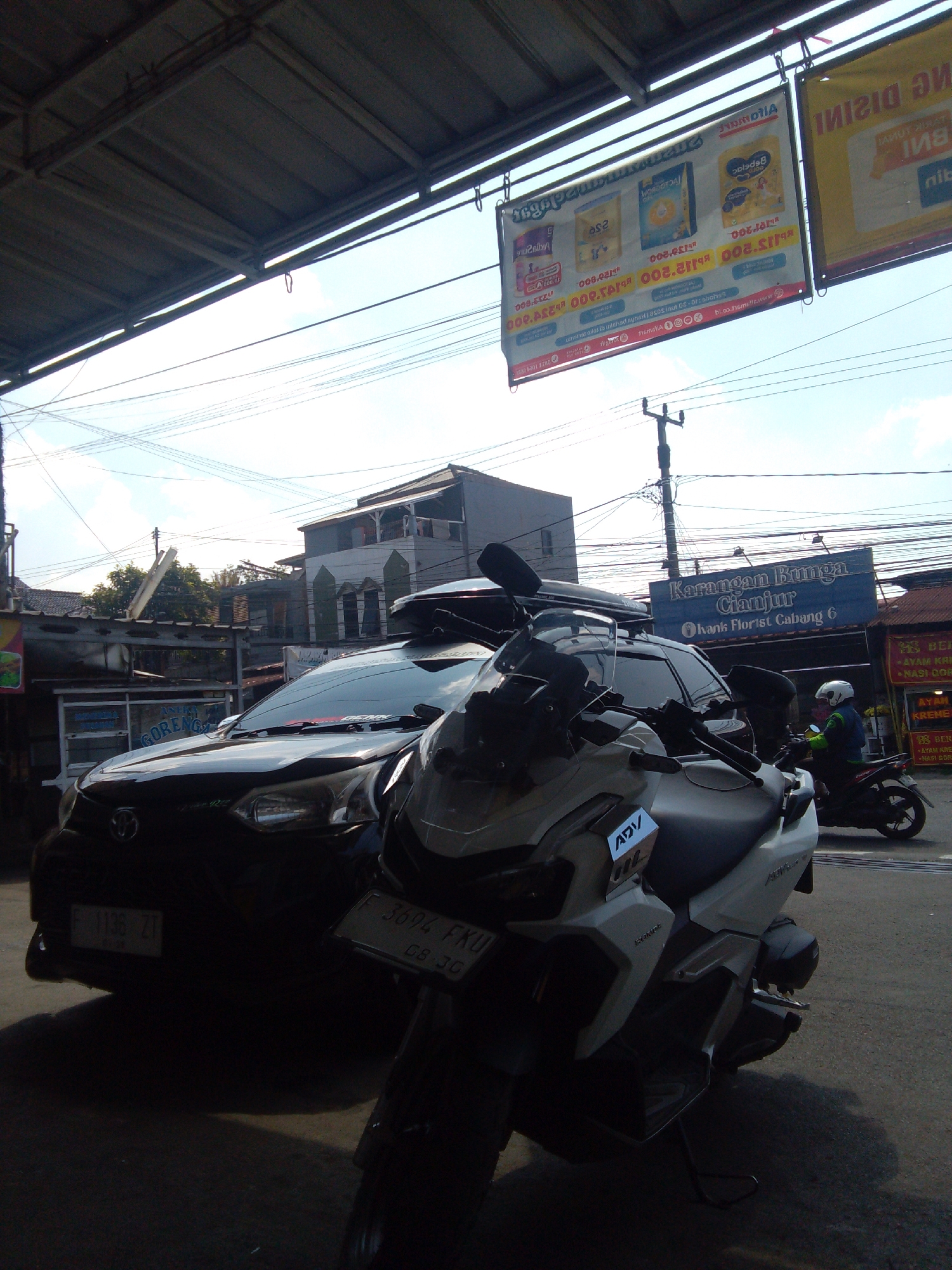 The image depicts a street scene in Cianjur, Indonesia. In the foreground, two motorcycles are parked. The motorcycle on the left is white and grey, with the "ADV" logo visible on its side. The motorcycle on the right is black and partially obscured by the white and grey motorcycle. Behind the motorcycles, a dark-colored car is parked, displaying a license plate that starts with "F 1136 ZT". Further in the background, a blue sign reads "Karangan Bunga Cianjur" which translates to "Flower Shop Cianjur," indicating the establishment's name and location. Several overhead power lines crisscross the sky. Buildings with distinct architecture are visible in the distance, and a person wearing a white helmet and a blue jacket is riding a black motorcycle away from the camera. The environment suggests an urban setting with commercial establishments and residential structures. The lighting indicates it is daytime, possibly late afternoon due to the shadows.