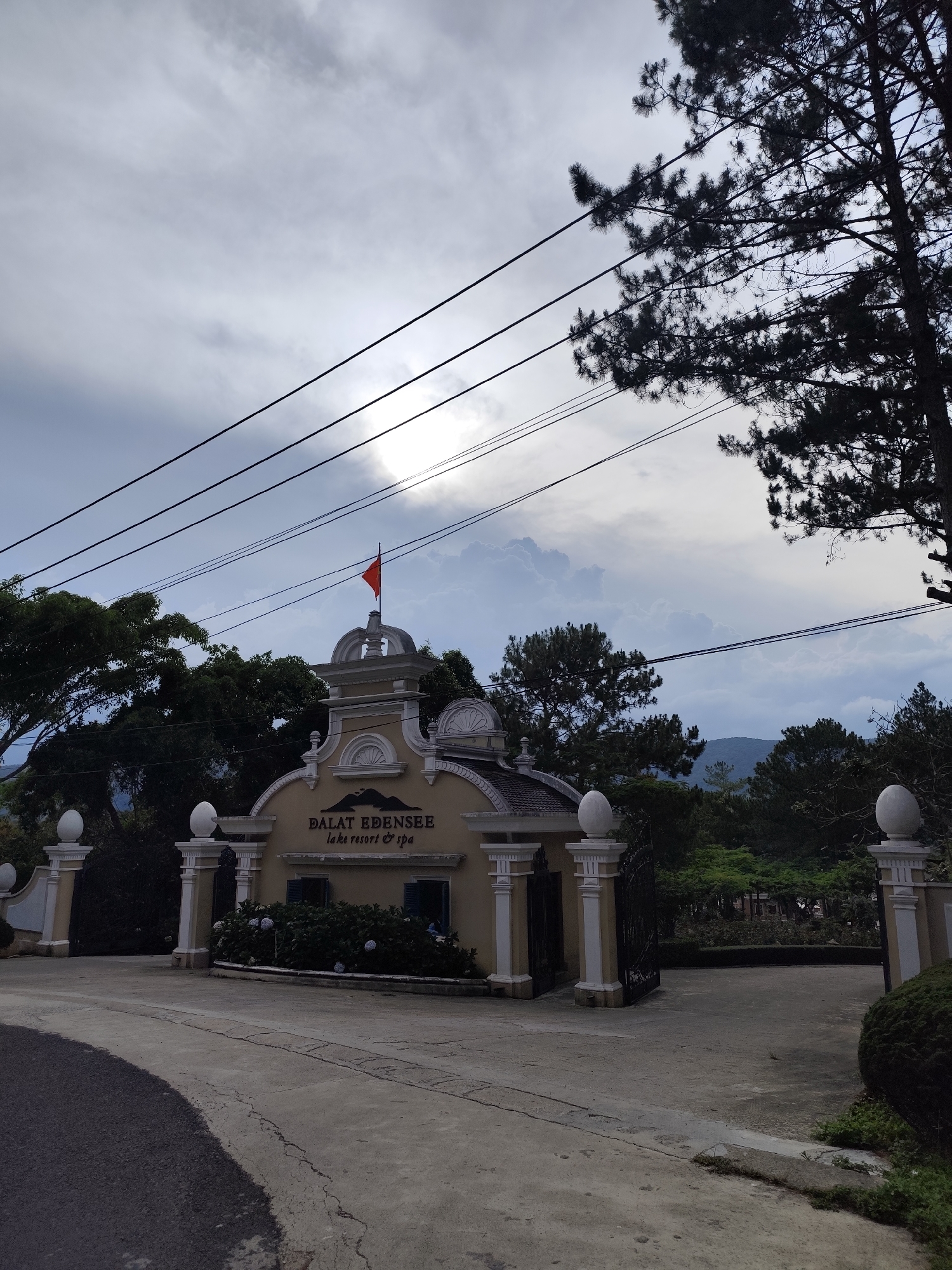 The image depicts the entrance gate of "Dalat Edensee Lake Resort & Spa", located in Da Lat, Vietnam. The gate is constructed of beige-colored material, featuring white pillars topped with spherical ornaments on either side. A small structure sits atop the gate, adorned with a red flag. Below the flag structure, the name of the resort is displayed.
The gate is surrounded by lush greenery and trees. A paved road leads to the gate. Overheads are electrical wires. The sky is cloudy, with some sunlight filtering through the clouds. There are no people visible in the image.