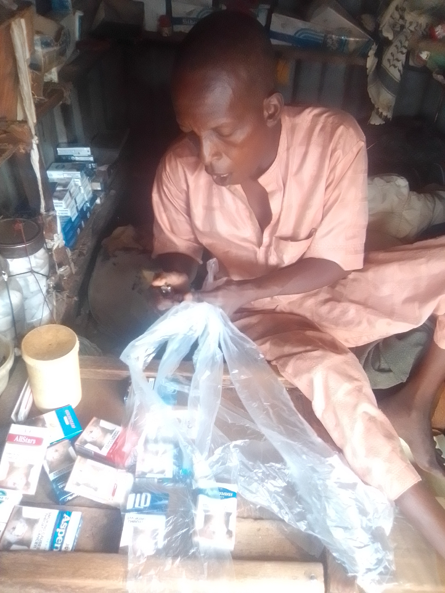 A man is seated in a dimly lit, cluttered stall, likely a small shop, in Pantame, Nigeria. He is wearing a light pink, loose-fitting garment. His gaze is directed downwards towards a collection of items laid out on a wooden surface in front of him. The items appear to be cigarette packs and possibly other small goods, some of which are partially obscured by plastic bags. The stall is filled with various objects, suggesting it is a place of business. The background is dark and indistinct, with shelves and more items stacked behind the man. The overall impression is one of a busy, perhaps modest, retail environment.