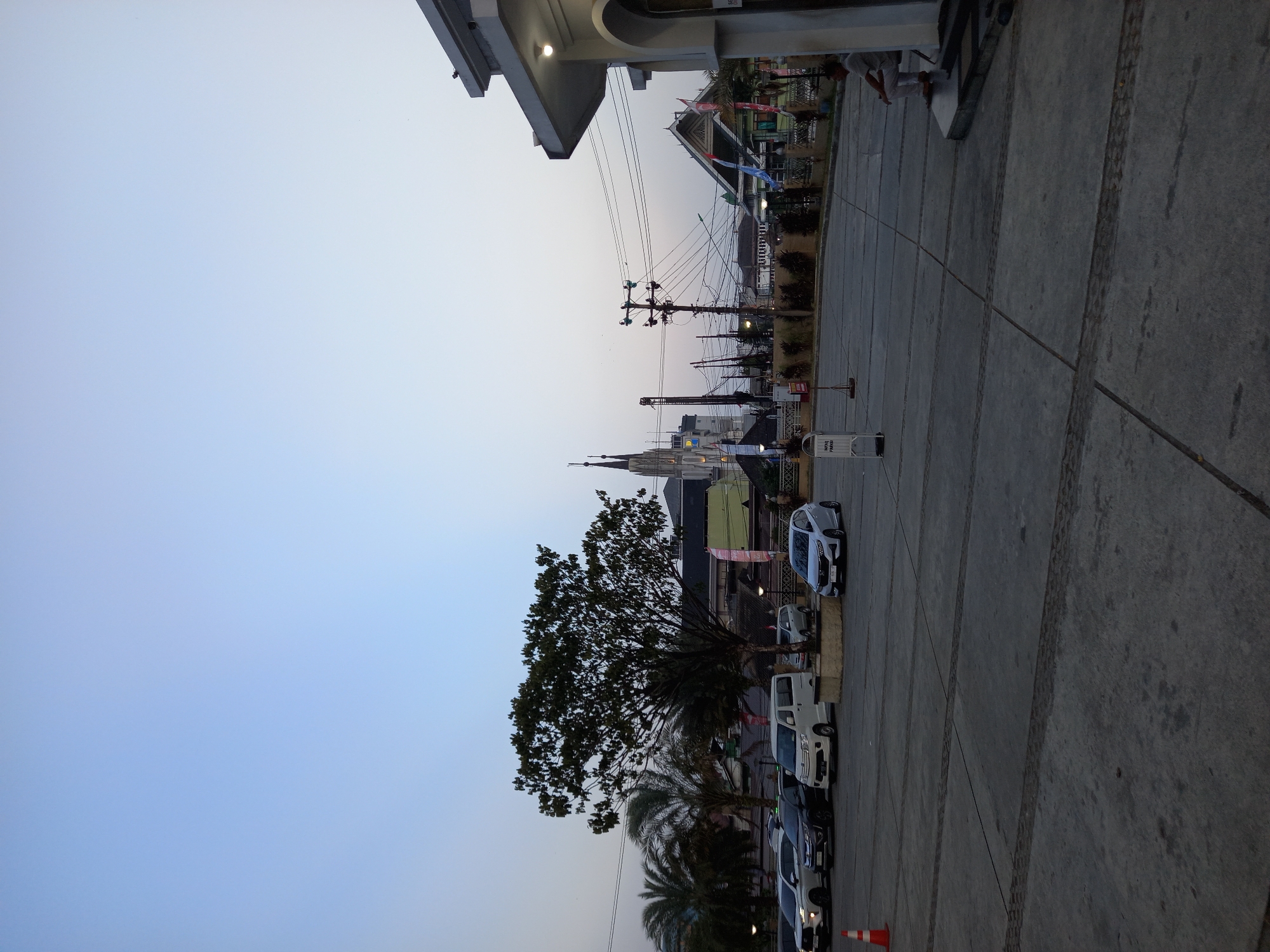 The image depicts an outdoor scene at dusk in Samarinda, Indonesia. In the foreground, a concrete parking lot is visible, with tire tracks marking the surface. A few cars are parked on the lot, including a white van, a white sedan, and other vehicles. To the left, a tall tree with dense foliage stands prominently. In the background, a church with spires is visible, along with several other buildings. Power lines and utility poles crisscross the sky. A man is seated on a step in the lower right portion of the image. The sky is a pale blue, indicating twilight.