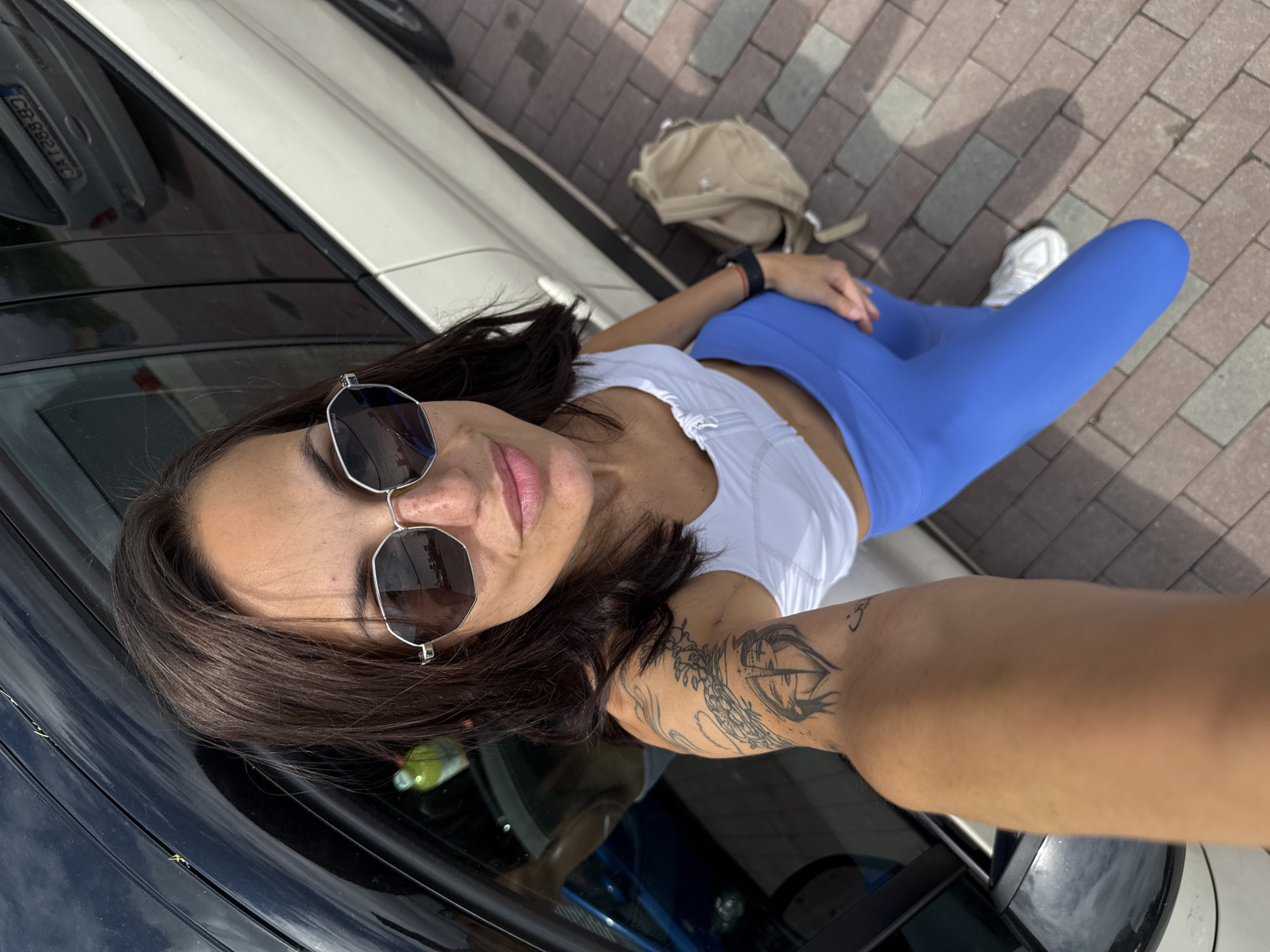 A woman wearing sunglasses, a white top, and blue leggings is positioned next to a car with the license plate CB 8821 AC, possibly in Pleven, Bulgaria. She has dark hair and a tattoo on her right arm. A light-colored backpack is on the ground near her. The surface under her feet is paved with bricks.