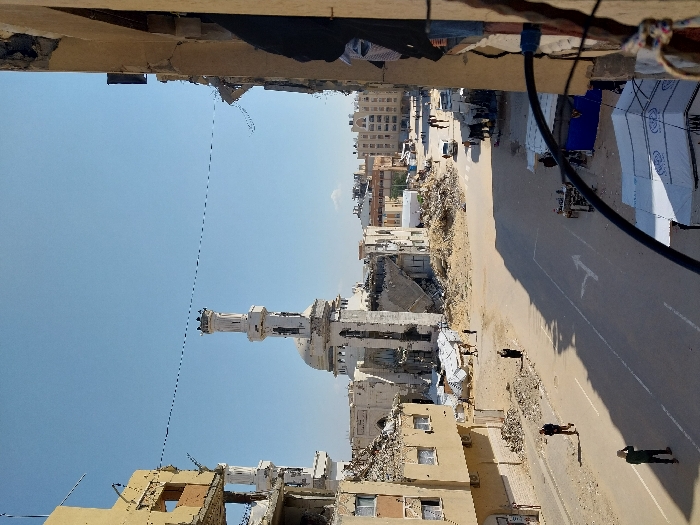 An elevated, wide-angle view captures a severely damaged urban street scene in Khan Yunis, Palestinian Territory, under a clear blue sky.
In the center-left background, a prominent mosque stands, exhibiting significant structural damage to its main body, though its tall, white minaret remains largely intact. To the immediate left, a multi-story building is extensively destroyed, with large sections of its facade missing, revealing interior spaces and exposed rebar. Further along the left side of the street, buildings are reduced to rubble or show severe damage. The perspective is from a damaged building on the right, with a partial view of its wall, a window frame, and objects resembling clothing hanging in the immediate foreground.
A paved road bisects the image, featuring a white arrow road marking indicating a right turn. The road itself appears cleared but is flanked by extensive piles of concrete rubble, dirt, and twisted metal from collapsed structures.
Approximately 10-15 individuals are observed walking within the scene. In the left foreground, two men walk along the debris-laden roadside. Another individual in dark attire is seen closer to the rubble on the left. On the right side of the road, several temporary structures, including white canopies (some appearing to be relief tents) and a blue tarpaulin tent, are set up. A white vehicle is visible on the road in the mid-distance on the right, with other vehicles further down the street. People are also observed walking in the distance along the street.
The scene is illuminated by bright daylight, casting distinct shadows from buildings and individuals. A dark cable or pipe extends into the foreground from the right side of the frame.