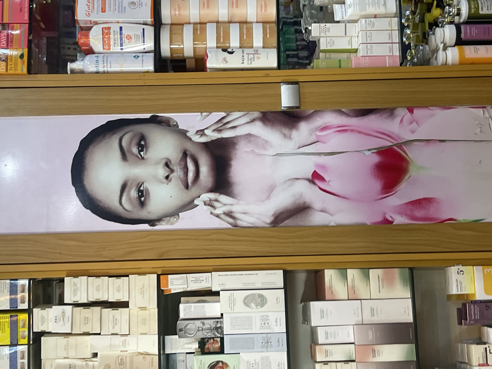 The image depicts a shelf in what appears to be a retail store, possibly in Galadimawa Village, Nigeria. The shelves are stocked with various skincare and cosmetic products. A promotional poster featuring a woman's face and a flower is positioned between two of the shelves. The products on the top shelf include brands like Gluta-C, Retin-A, and Kojie-san, while the lower shelf contains a range of skincare products packaged in white and cream-colored boxes with minimal labeling. The focus is on the display and arrangement of these products within the store environment.