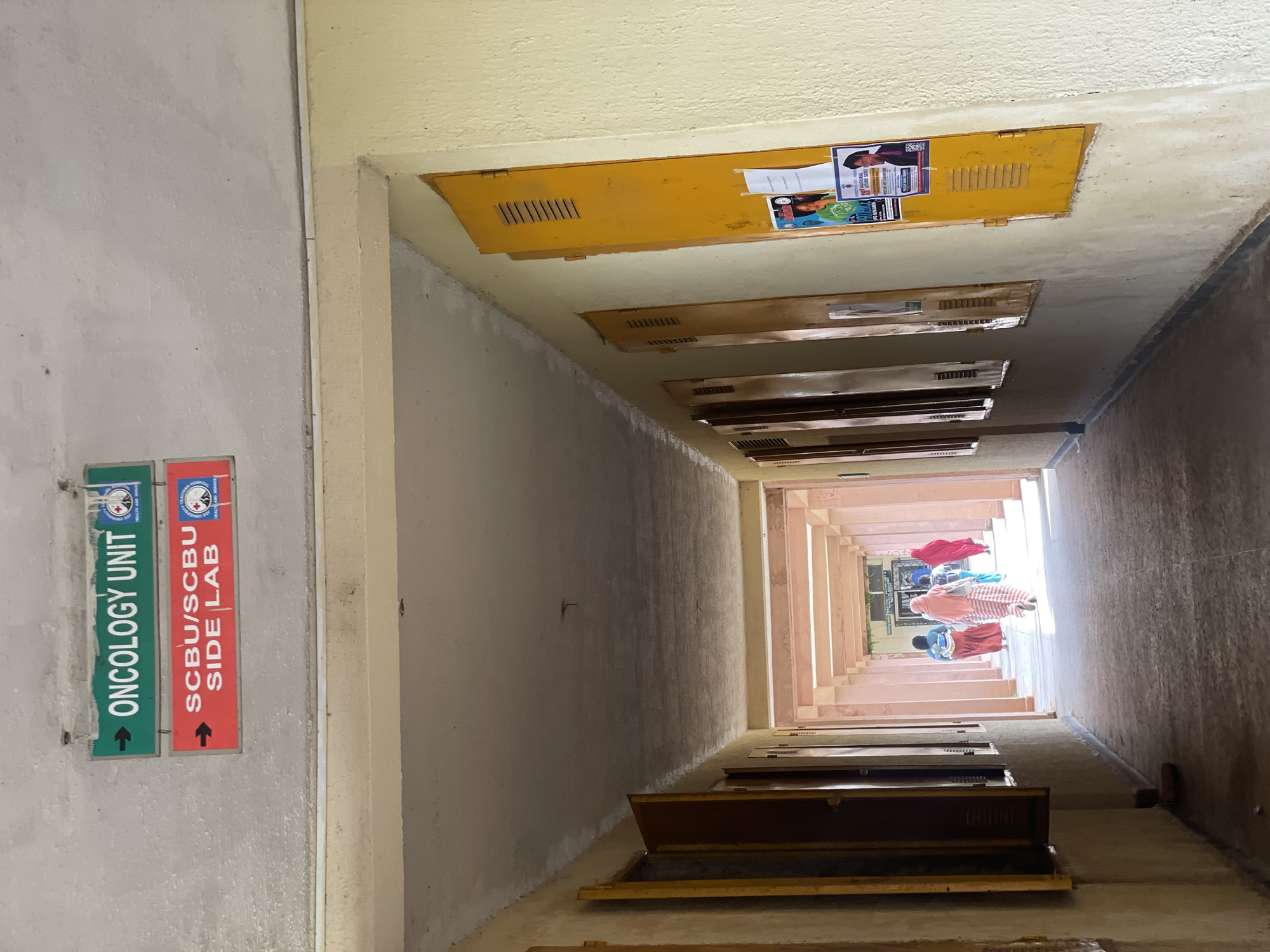 The image shows a hallway with two signs overhead: "ONCOLOGY UNIT" in green and "SCBU/SCBU SIDE LAB" in red. The hallway has a concrete floor and a light-colored ceiling. On the right side of the hallway, there are yellow lockers with some posters attached to the wall. On the left side, there are windows with open shutters and a view of another hallway with people walking. The hallway appears to be part of a hospital or medical facility. The location is specified as Gwong, Nigeria.