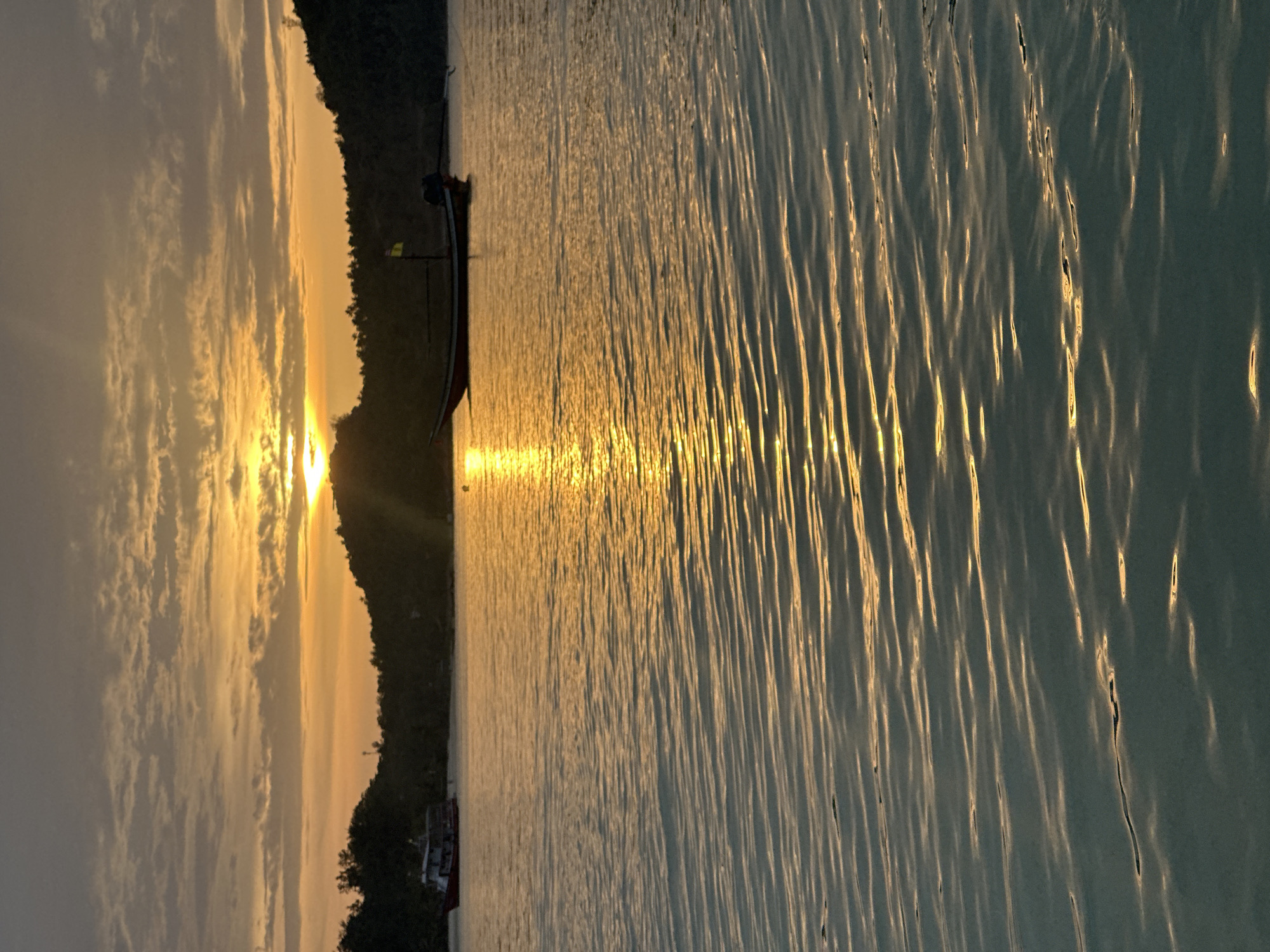 The image depicts a serene sunset over the ocean in Ko Pha-ngan District, Thailand. The sky is illuminated with warm hues of orange and yellow, with scattered clouds catching the sunlight. Two boats are visible in the water. One is a smaller, red and white vessel in the distance to the left. Closer to the center and right, a longer, narrow boat with a yellow flag is anchored. The ocean surface is calm, with gentle ripples reflecting the golden light of the setting sun, creating a shimmering effect. The background features a silhouetted treeline of a hilly island.