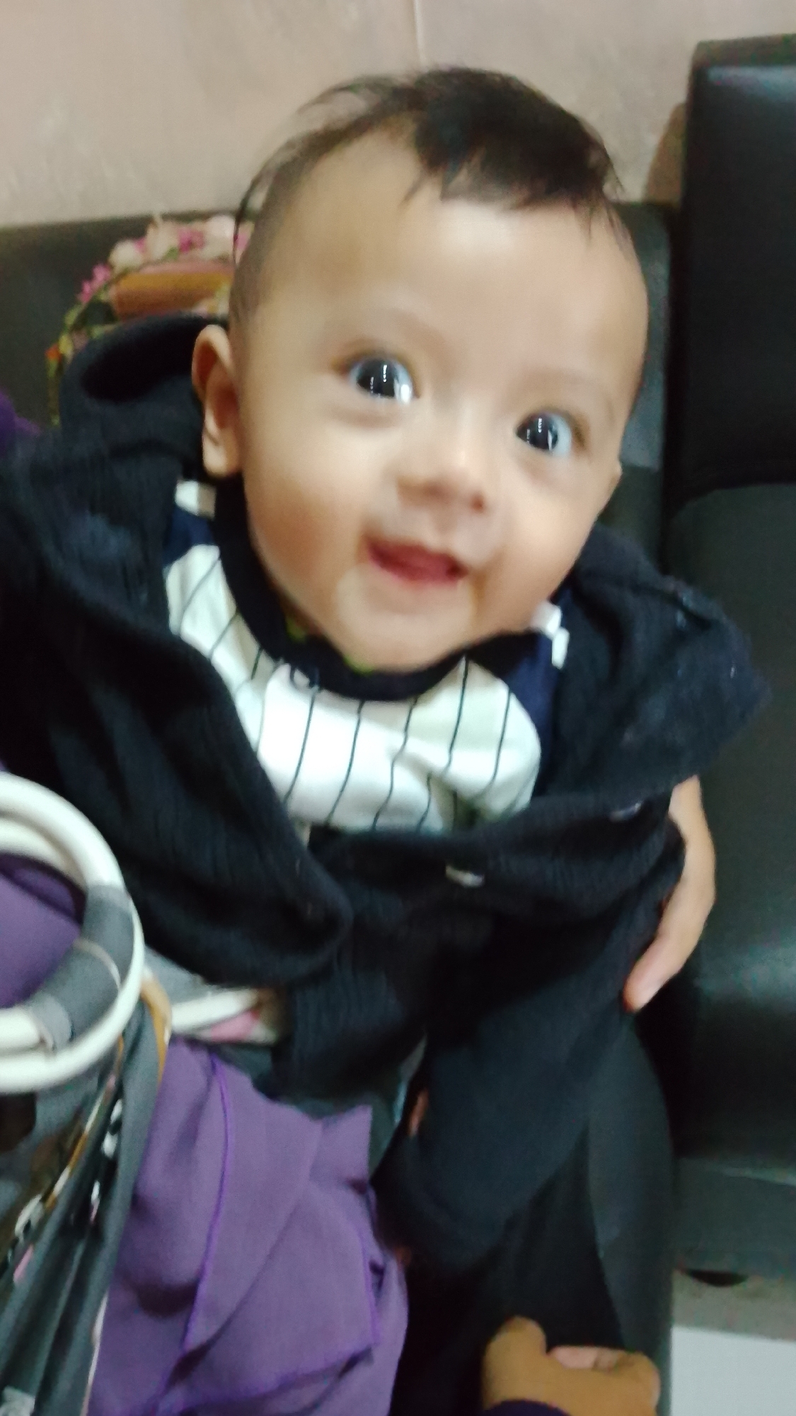 A close-up, waist-up shot captures a baby with fair skin, dark hair, and wide, dark eyes, looking directly at the camera. The baby is wearing a black hooded jacket over a white shirt with dark blue pinstripes. The baby's left arm is visible, and their right arm is mostly obscured by the jacket. The baby is being held by an unseen person, with a hand and forearm visible on the right side of the frame. In the lower left corner, a portion of a purple fabric is visible, potentially part of a garment or blanket. The background is blurred, with a dark surface, possibly a couch, and a lighter wall. The lighting is dim, suggesting an indoor setting, likely a residence in Bojongsoang, Indonesia.