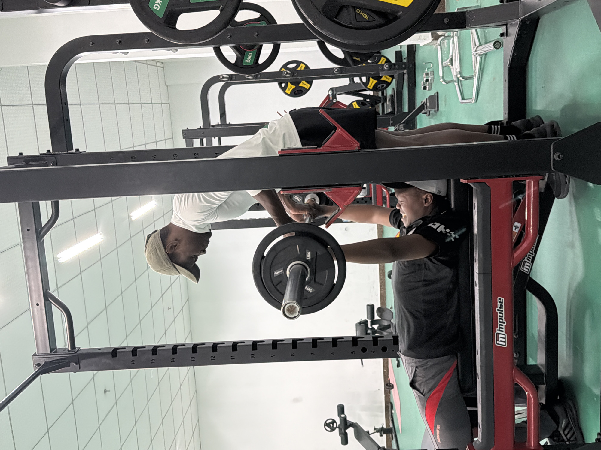 The image, taken upside down, shows two men in a gym. The man on the left, seen from the waist up, wears a white t-shirt and a baseball cap. He is adding weight to a barbell. The man on the right, from the waist up, wears a black and grey shirt and a grey cap. He assists in adding the weight. The barbell sits on a weight rack marked with numbers "16" through "7." Weights are stored on the rack above them. To the right of the assisting man, the gym equipment has the word "impulse" on it. The gym is located in Jiangning District, China.