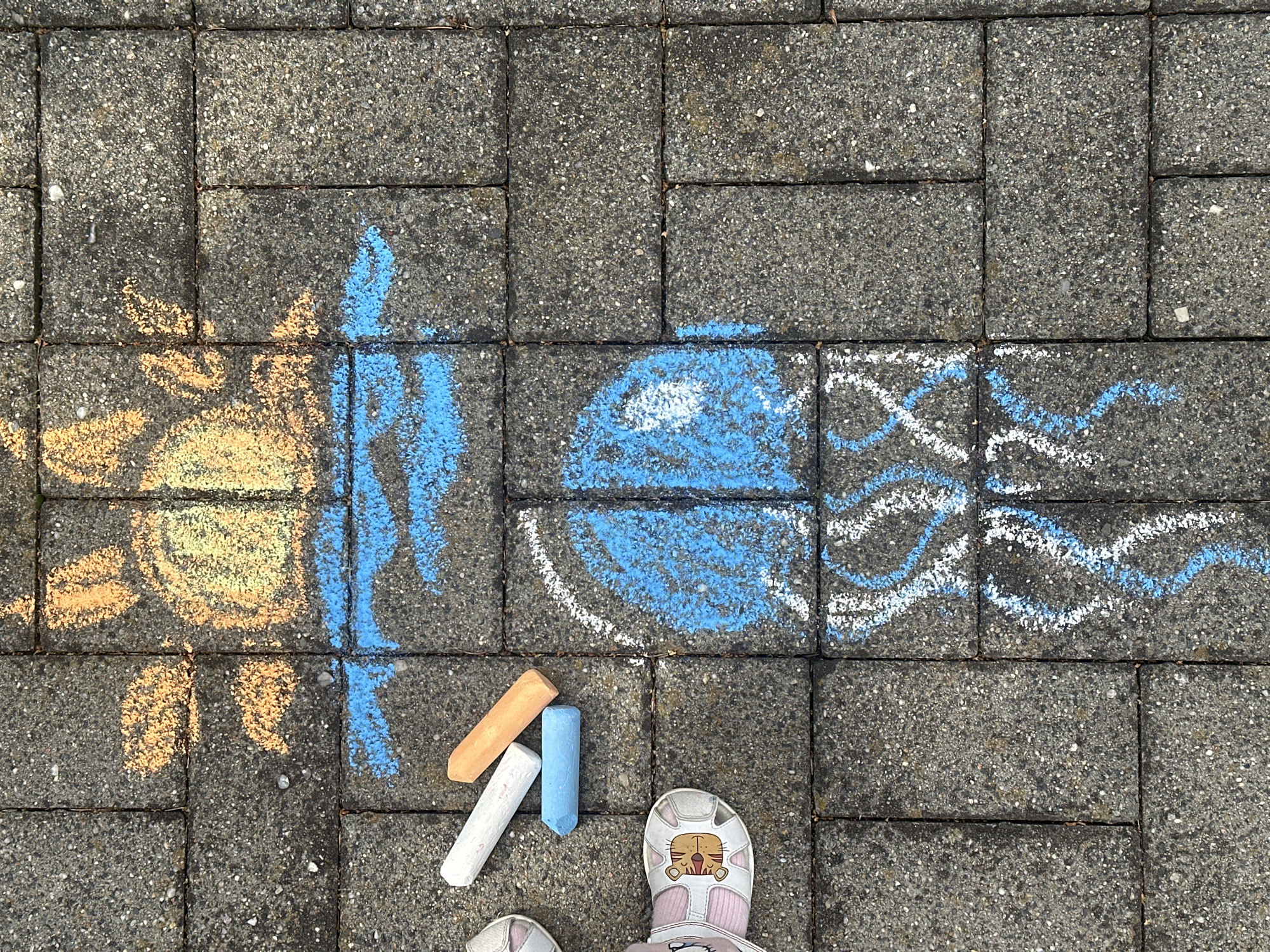 The image depicts a chalk drawing of a sun over water, with a jellyfish beneath, rendered on a paved surface. Three sticks of chalk, one white, one orange, and one blue, are positioned to the left of the drawing. A child's pink sandal, adorned with a tiger face, is visible in the bottom left corner, along with a portion of another white sandal. The drawing is on a grey, textured paved surface, likely cobblestones or similar paving blocks. The drawing itself features a yellow and orange sun with rays, blue wavy lines representing water, a large blue jellyfish body with white highlights, and wavy blue and white tentacles. The scene appears to be outdoors, and the paving suggests a sidewalk or patio.