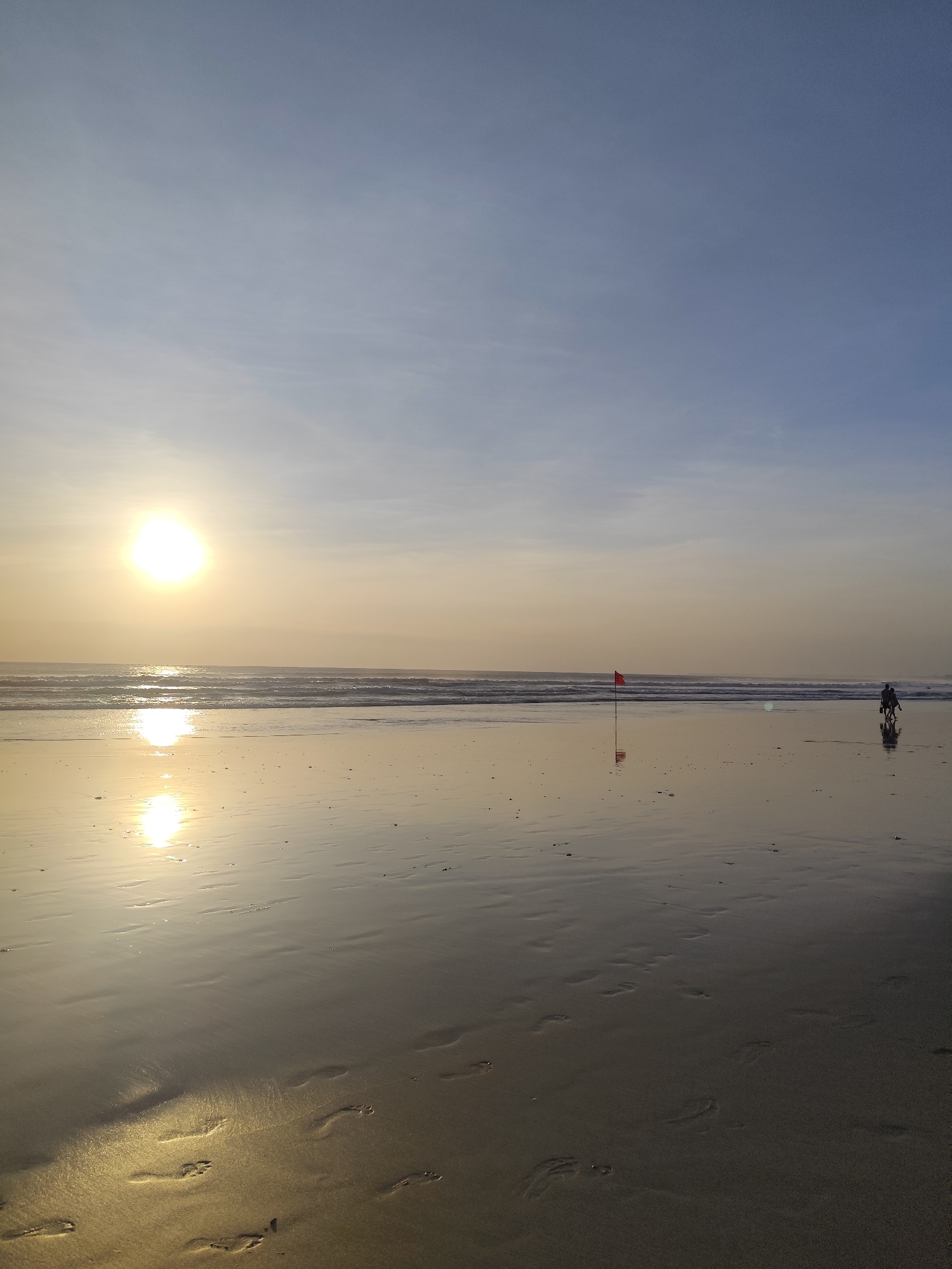 An image taken at eye level shows a beach at sunset. The sun is low in the sky, casting a warm glow across the scene. The ocean is calm, with gentle waves lapping at the shore. Footprints are visible in the wet sand, indicating recent human activity. A red flag stands on the beach, possibly marking a hazard or a boundary. In the distance, two figures can be seen walking along the shoreline, silhouetted against the setting sun. The sky is a pale blue, with wispy clouds stretching across the horizon. The overall mood of the image is peaceful and serene, evoking a sense of tranquility and natural beauty.