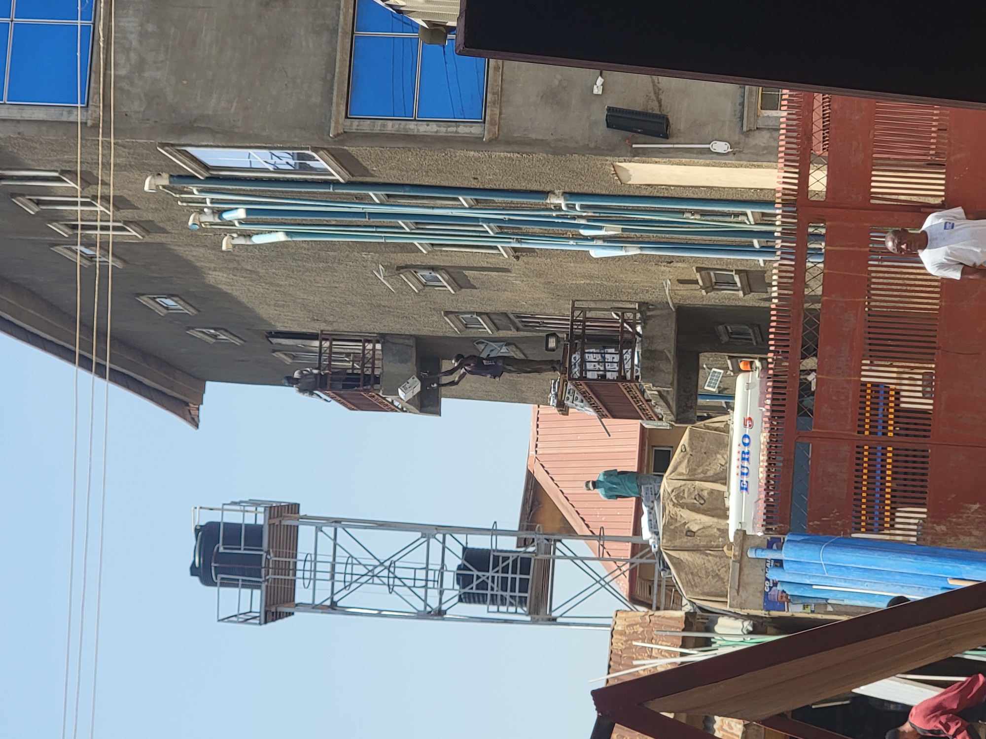 The image depicts an outdoor scene in Maiburiji, Nigeria. A multi-story concrete building with blue-framed windows and numerous pipes running along its exterior is visible. In the foreground, a metal water tower with two black tanks stands against a clear blue sky. Adjacent to the building, a partially visible truck with the word "EURO" displayed vertically is parked near a stack of blue pipes. A red metal gate and fence separate the scene from a figure in a white shirt standing nearby. Several individuals are present, some obscured by buildings and structures. The environment includes buildings with reddish-brown roofs and a mix of residential and commercial structures, giving the impression of a built-up urban area.