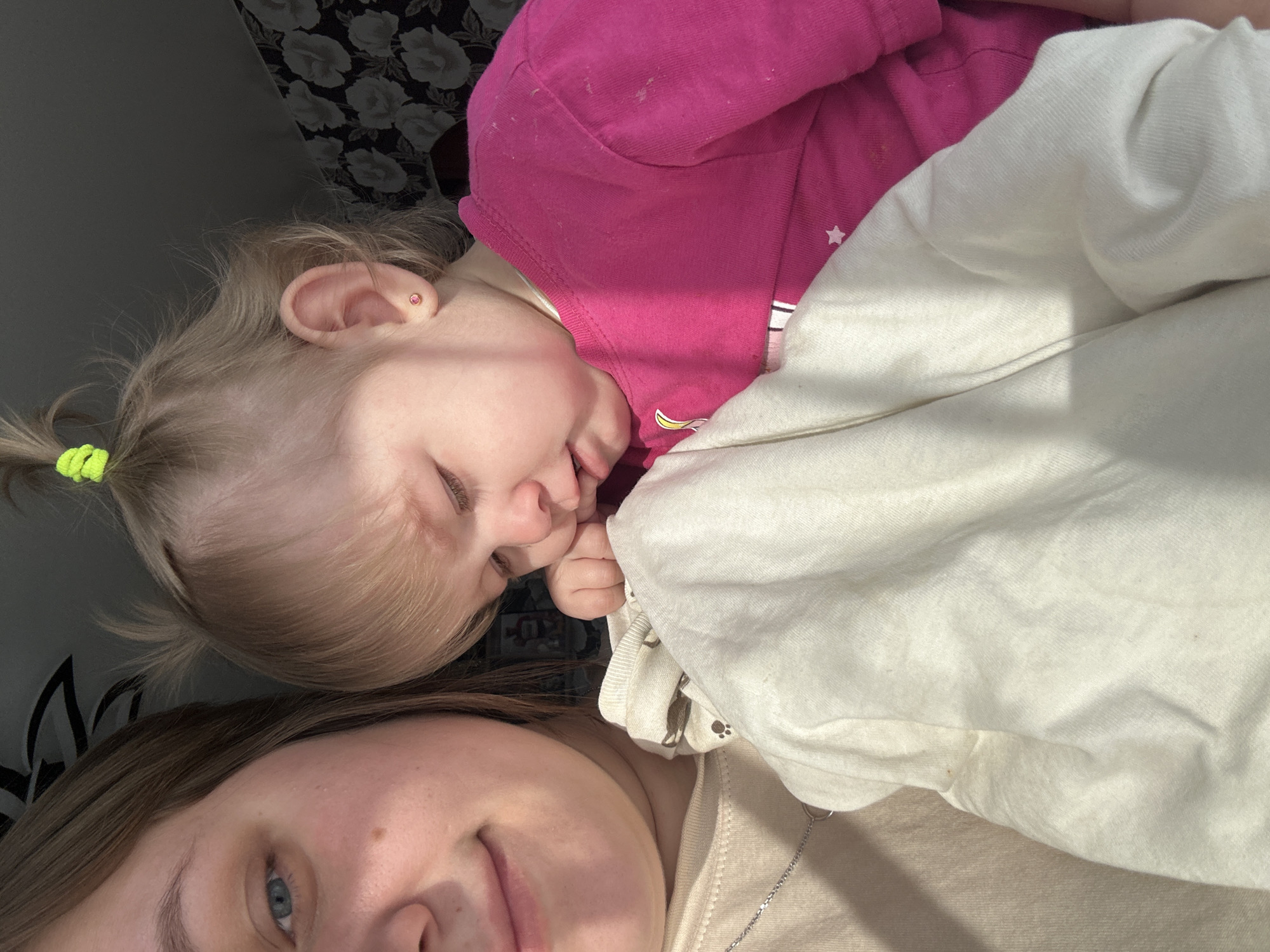 A young woman and a toddler lie next to each other, appearing to be resting or sleeping. The woman has light brown hair and blue eyes, and is wearing a beige top with a silver necklace. The toddler has blonde hair pulled back in a small ponytail with a green hair tie, and is wearing a pink shirt. The background shows a floral pattern on what appears to be a couch or bed. The location is not discernible from the image and cannot be determined to be in Ольховка, Russia.