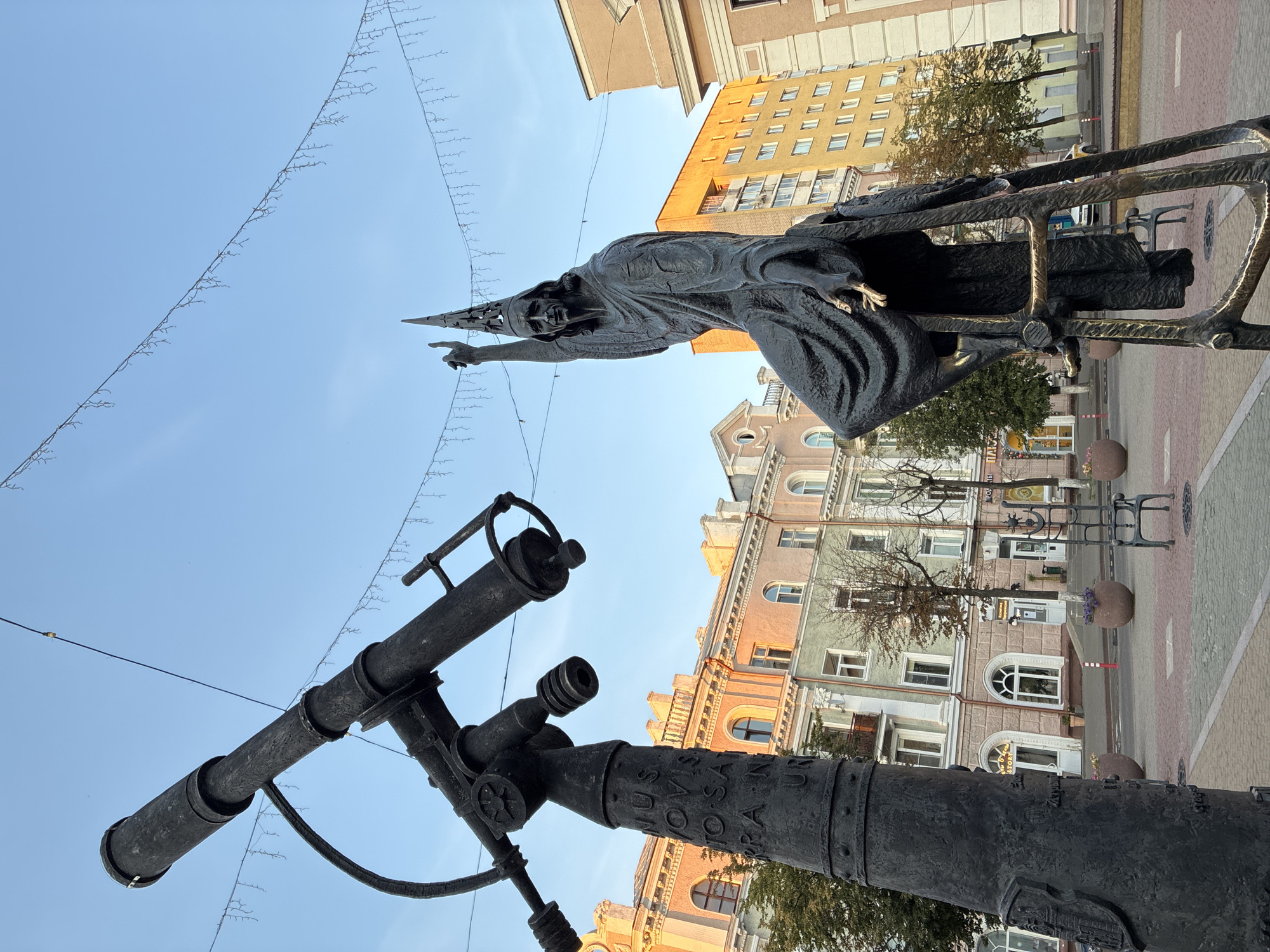 The image depicts a bronze sculpture of a man in a conical hat and flowing robes, with his right arm extended upwards, pointing. He is seated on a metallic structure resembling a ladder or scaffolding. To the left of the sculpture is a large bronze telescope mounted on a pedestal. The telescope's pedestal is inscribed with text. In the background, there are multi-story buildings with light-colored facades, some featuring arched windows. Trees and decorative string lights are also visible. The scene appears to be in a public square or street, paved with brickwork. This sculpture is located in the city of Mahilyow, Belarus, and is a monument to the Belarusian astronomer and physicist Josip Zhyzhefsky.