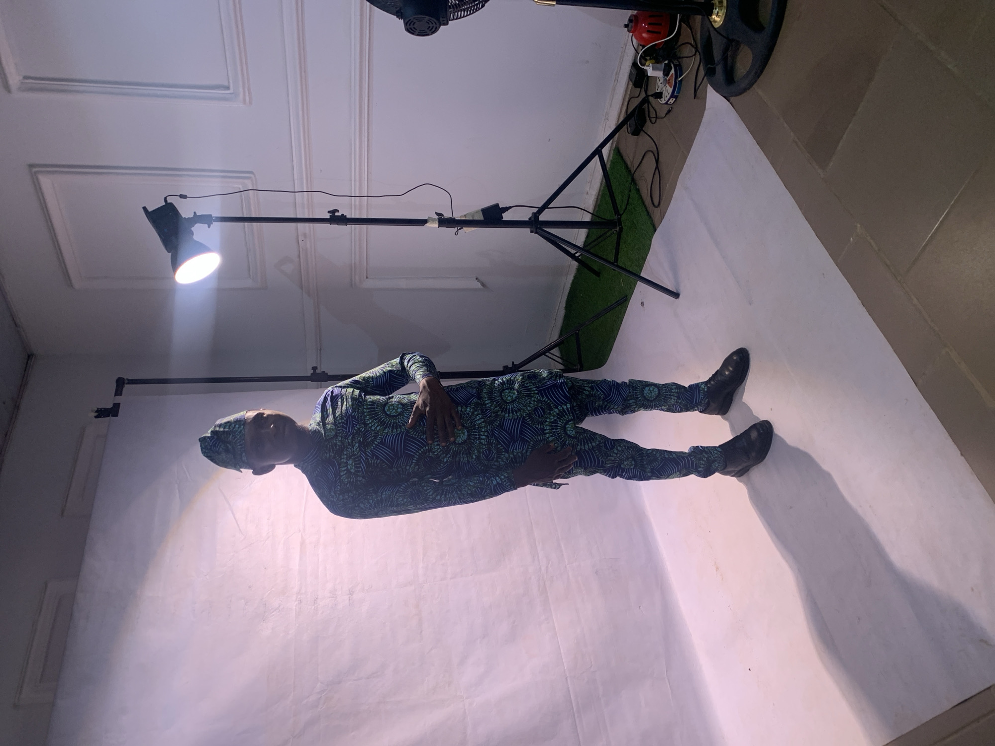 The image shows a man in a dark blue and green patterned suit standing on a white backdrop. The backdrop is illuminated by a bright studio light on a stand. An oscillating fan and a power strip are visible behind the light stand. The scene appears to be indoors, possibly a makeshift studio setup. The setting, inferred from the prompt, is Jos, Nigeria.