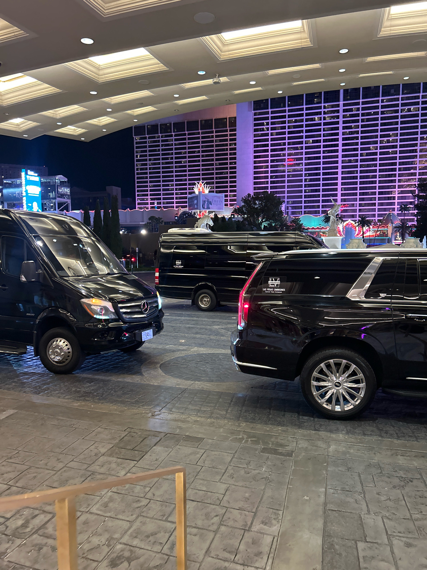 The image depicts a nighttime scene in front of a hotel in Paradise, Nevada, United States of America. Three black luxury vehicles are parked on a cobblestone-like pavement. In the foreground, a Mercedes-Benz Sprinter van is partially visible on the left. Behind it, another black Mercedes-Benz Sprinter van is parked. To the right, the rear section of a black Cadillac Escalade is prominent, displaying a "LAS VEGAS LIMOUSINES" sticker. In the background, a tall, modern hotel building is illuminated with purple and pink lights, featuring a grid-like pattern of windows. The sky is dark, indicating nighttime. A walkway with a golden railing is visible in the bottom left corner.