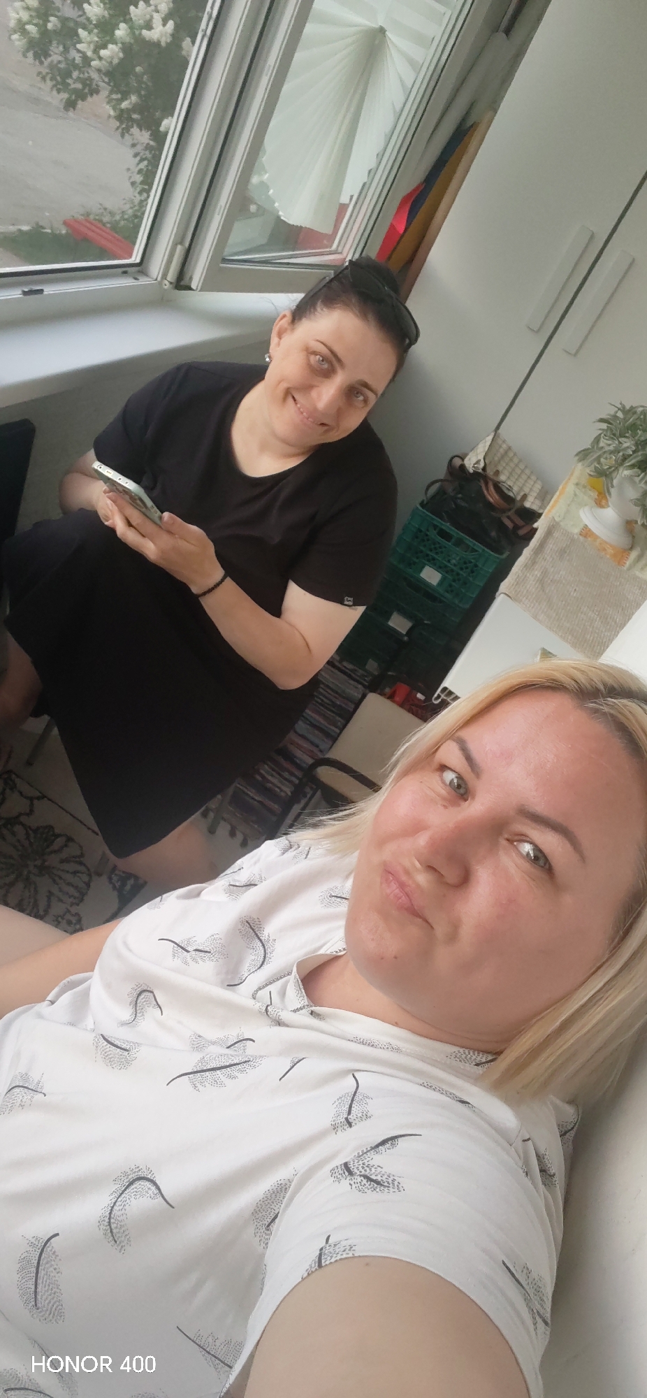 Two women are indoors. One is fair-haired and wearing a white patterned shirt. She is closer to the camera. Behind her, another woman with dark hair, wearing a black dress, is looking at a phone. They appear to be in a room with a window. The text "HONOR 400" is in the lower left corner. The location is in the city of Noviy, Russia.