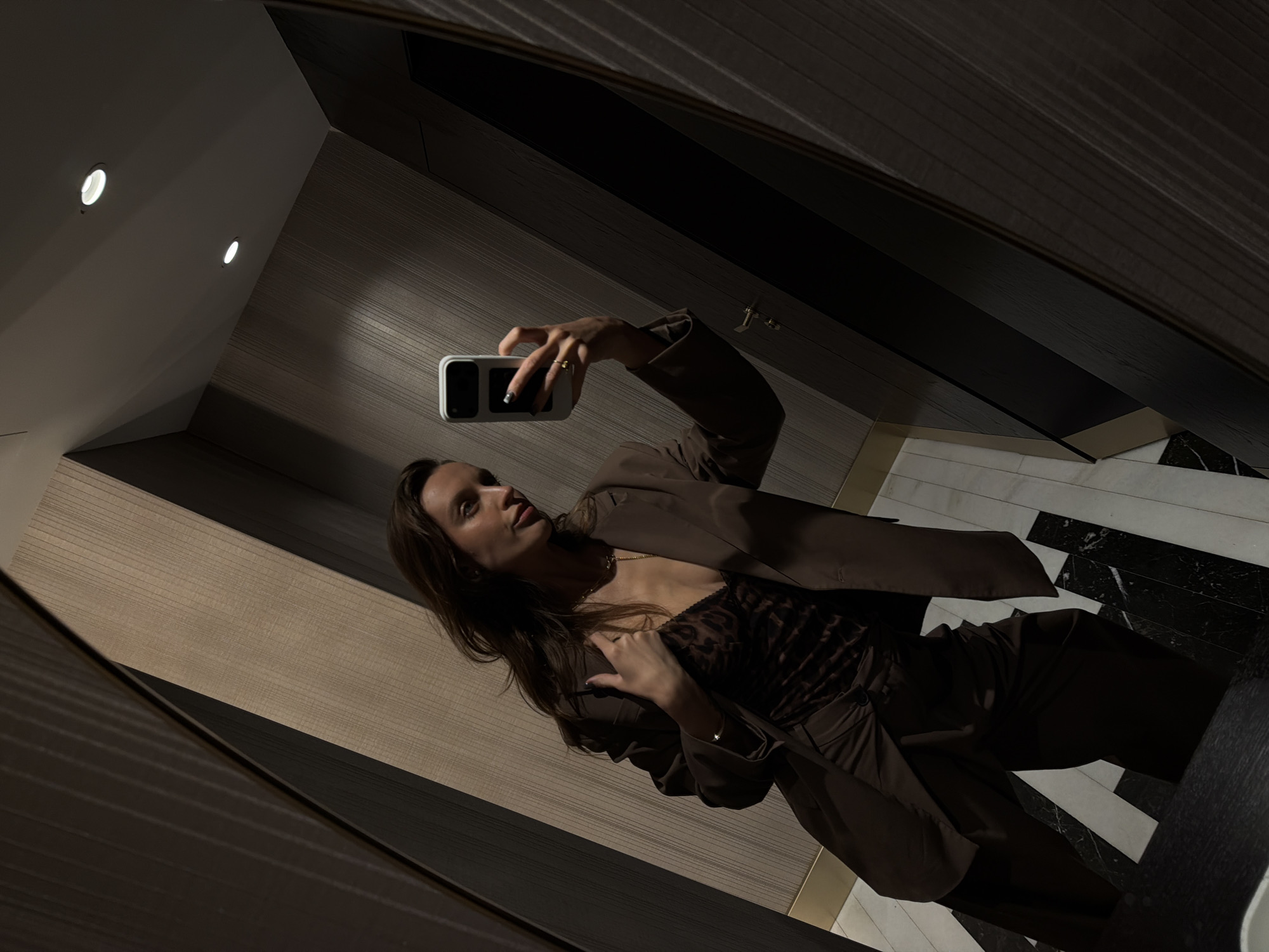 A woman is taking a mirror selfie in what appears to be a bathroom. She is wearing a brown suit over a patterned top. The bathroom features wood-paneled walls, recessed lighting, and black and white marble flooring. There is no information provided to confirm it is in Budva, Montenegro.