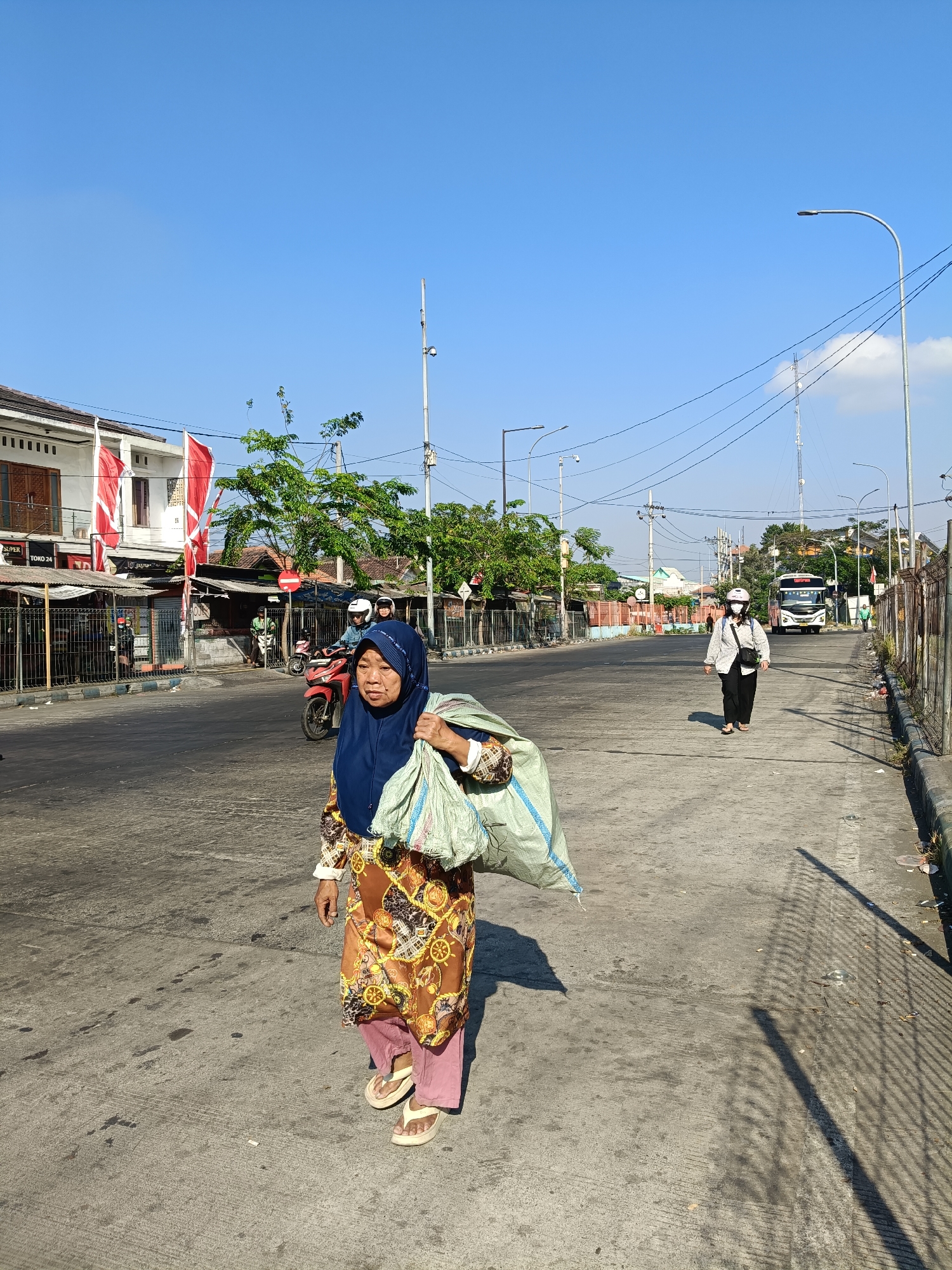 An Indonesian woman wearing a navy blue hijab and a patterned tunic walks on a street in Kedungrejo, Indonesia. She is carrying a large, light-colored bag over her shoulder. In the background, a person in a helmet and mask walks away from the camera, and a bus is visible on the road. The street is lined with buildings, trees, and utility poles. The sky is clear and blue.