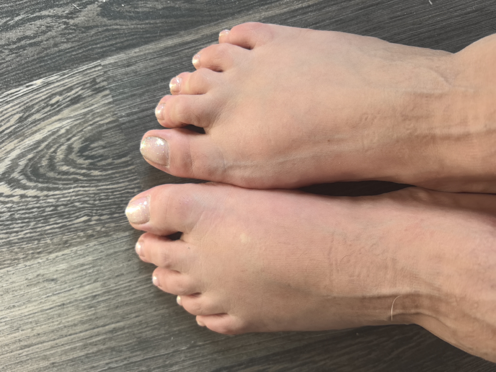 Two bare feet with lightly glittered toenails are positioned on a dark gray wooden floor. The feet are light-skinned and appear to be slightly swollen, with visible veins on the top of the left foot. The wooden floor has a distinct grain pattern, with planks running horizontally across the image. The lighting is natural, casting soft shadows. The image appears to be a close-up, taken from a slightly elevated angle. There are no other people or objects present. The location is not discernible from the image.
