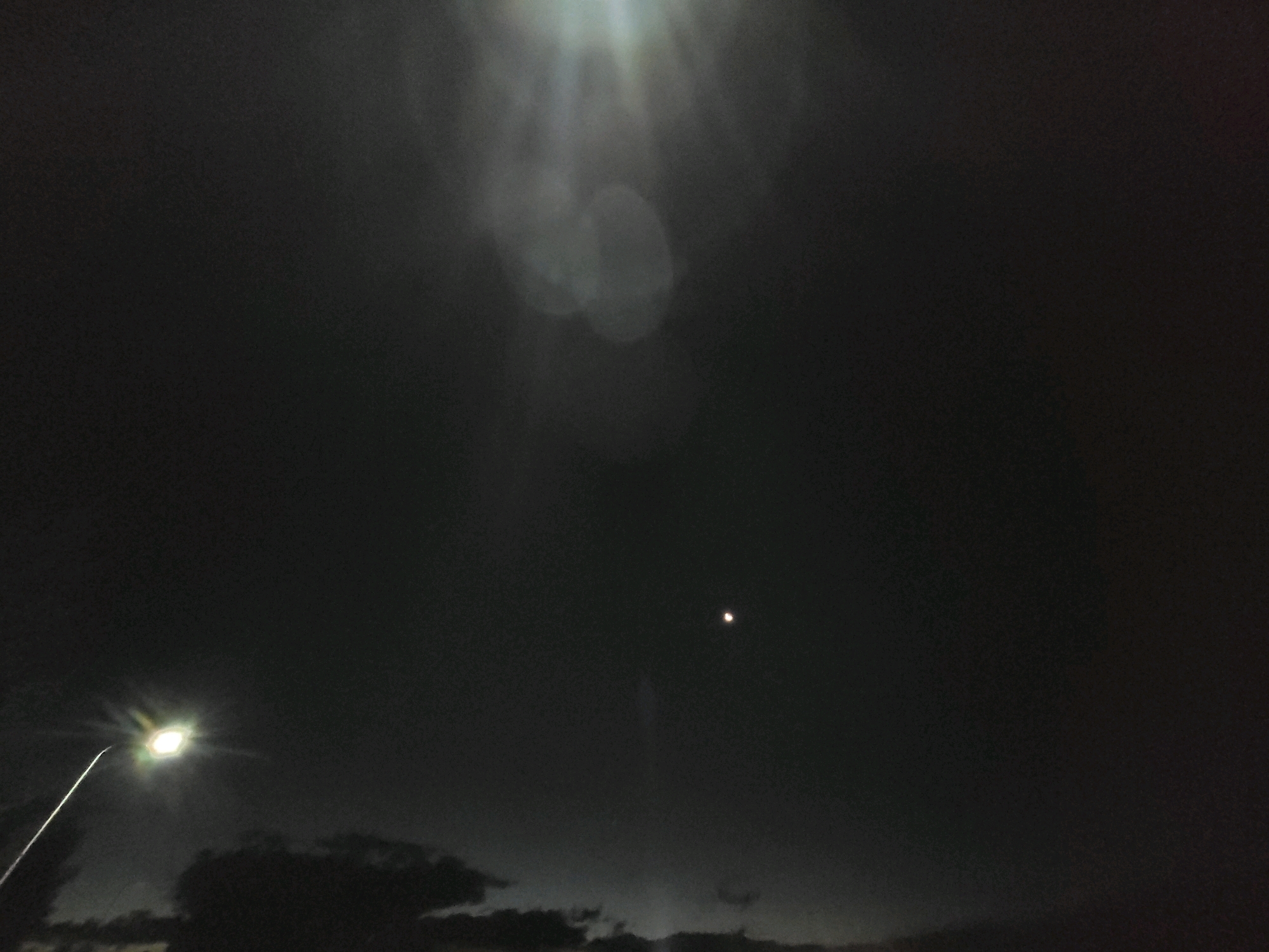 An outdoor nighttime photograph taken in Ozolnieki, Latvia, captures a dark sky with a bright street lamp on the left side of the frame. The street lamp is emitting a strong, diffused light that casts a glow into the surrounding darkness. In the upper portion of the image, several out-of-focus circular light orbs are visible, suggesting lens flare or atmospheric conditions. Towards the center-right of the frame, a small, bright celestial body, possibly a planet or star, is visible against the dark sky. The bottom of the image shows the silhouettes of clouds or trees against a slightly lighter horizon.