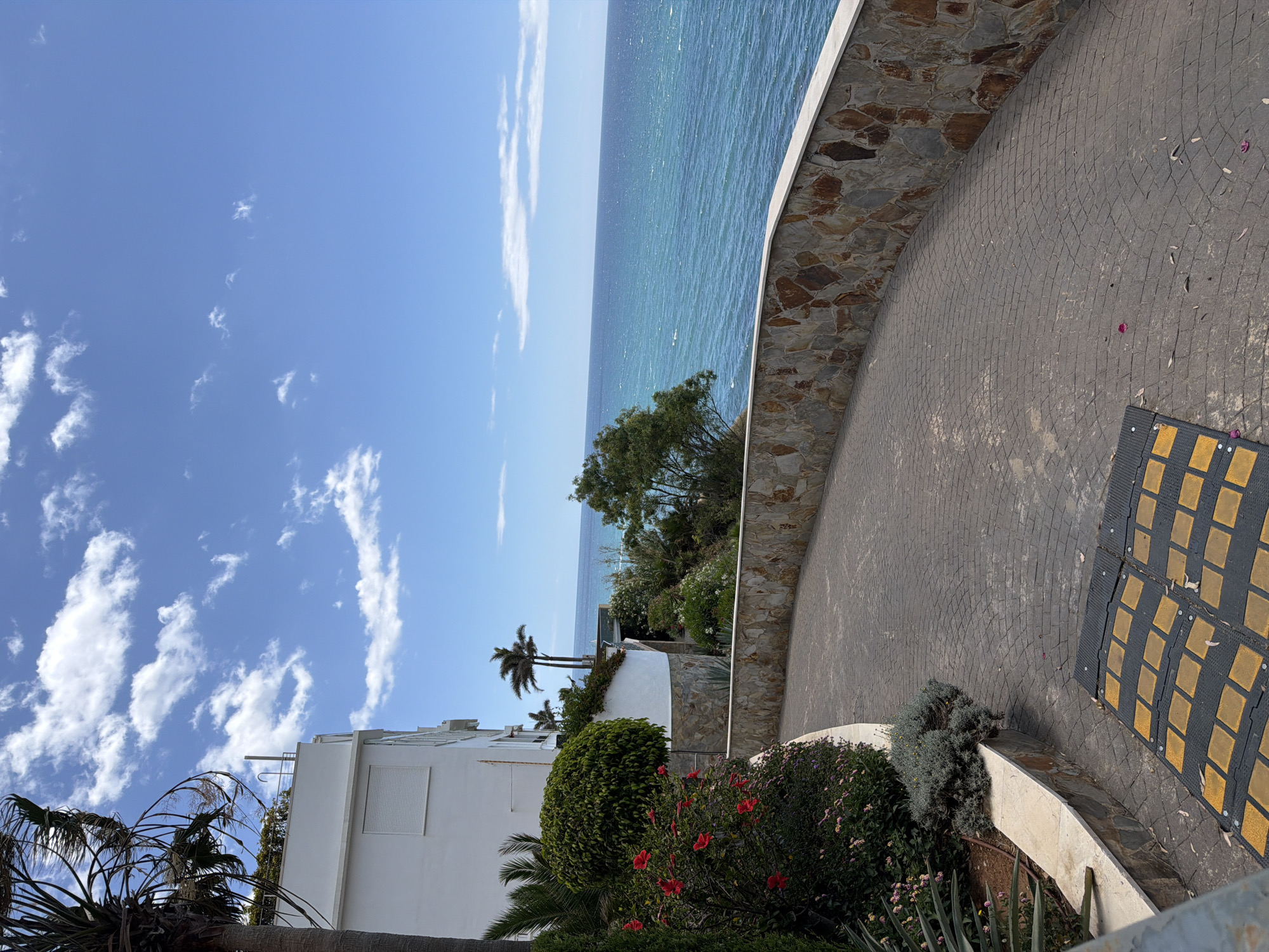 The image depicts a coastal scene in Benalmádena, Spain. The view is from an elevated position looking towards the Mediterranean Sea. A stone wall curves along a paved road, providing a barrier between the road and the vegetation. The sea is calm with a blue-green hue under a bright blue sky with scattered white clouds. A white building is partially visible on the left side of the frame. Palm trees and other shrubs are interspersed in the foreground. In the bottom right of the frame, there is a speed bump.