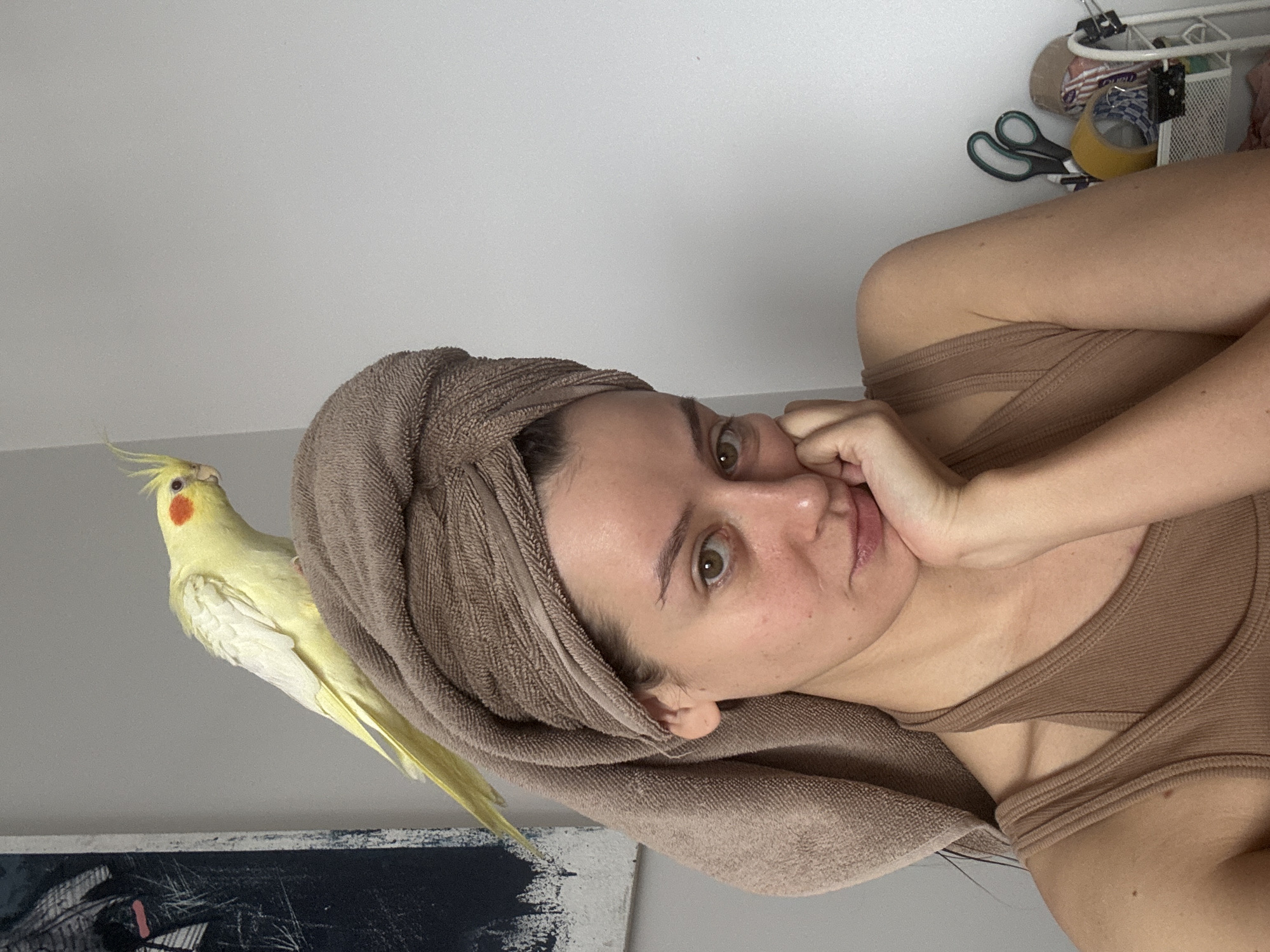 A woman with a towel wrapped around her head is holding her face in her hand and looking at the camera. A yellow cockatiel is perched on the edge of a painting next to the woman. Scissors and other items are visible on a shelf above the woman. The image appears to be taken indoors. There is no location in Krasnoyarsk, Russia, discernible from the image.