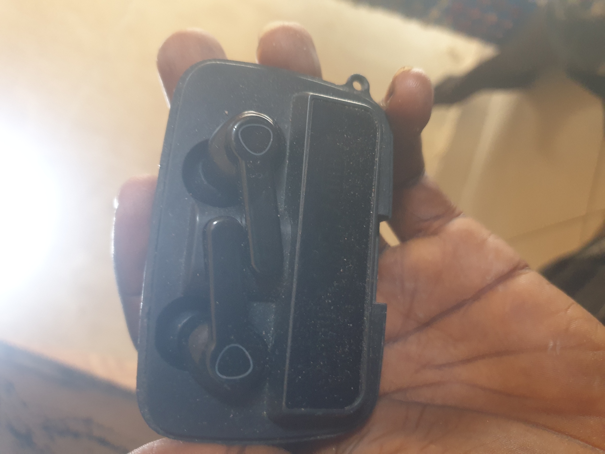 A close-up, top-down shot displays a black earbuds charging case, held in a person's hand. The case is open, revealing two black wireless earbuds nestled inside. The earbuds have a distinct oval shape with a metallic ring accent at the tip of each. The charging case itself is rectangular with rounded corners and a textured interior. The person's hand, with visible fingerprints and some dust particles, is the primary subject holding the case. The background is blurred, suggesting an indoor environment, possibly a room with natural light creating a bright, washed-out effect. The scene does not provide any specific visual cues to identify a location within Jalingo, Nigeria.