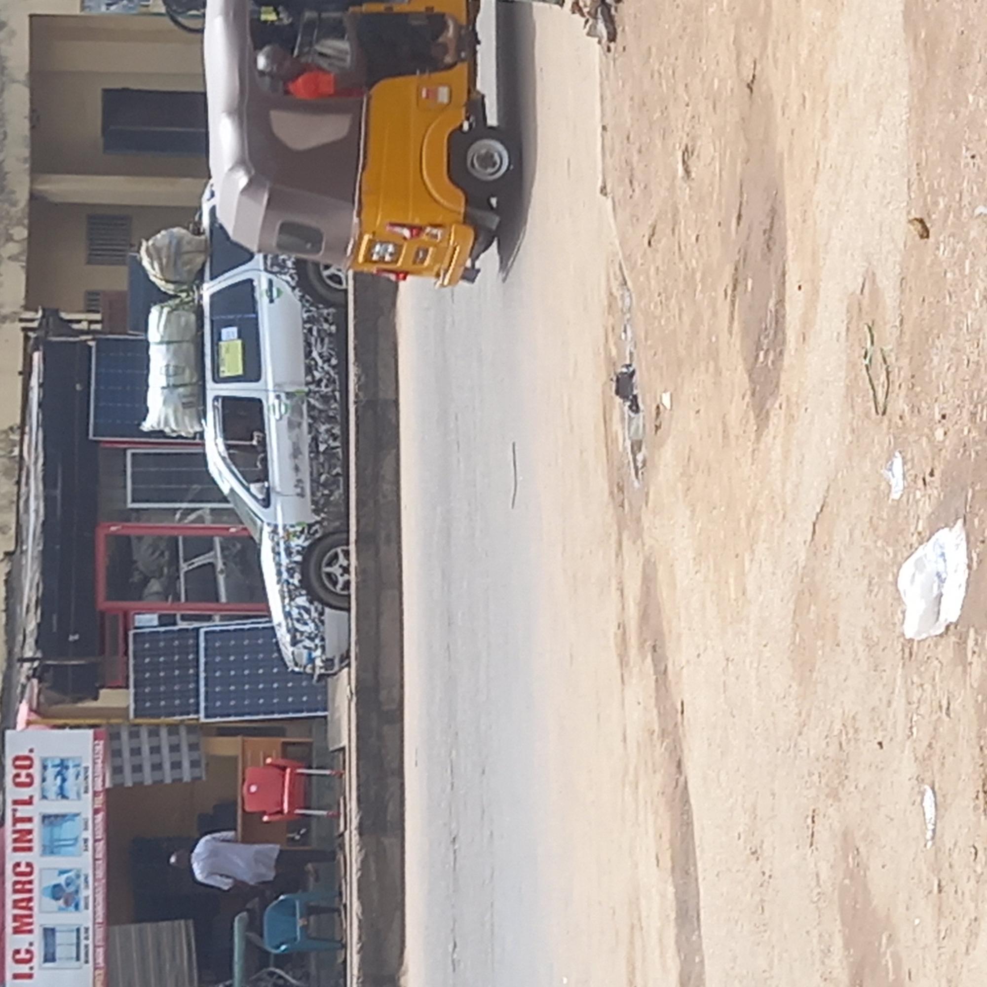 The image, taken in Dadi Riba, Nigeria, shows a street scene with a white car covered in camouflage, carrying bundled goods strapped to its roof, parked on the side of the road. A yellow three-wheeled vehicle is partially visible just behind the car. On the left side of the frame, a building with the signage "I.C. MARC INTL CO." is visible, displaying what appear to be household items. Solar panels are stacked against the building's facade. A man dressed in a white shirt is standing outside the building. The street itself appears to be paved, and the surrounding ground is sandy.