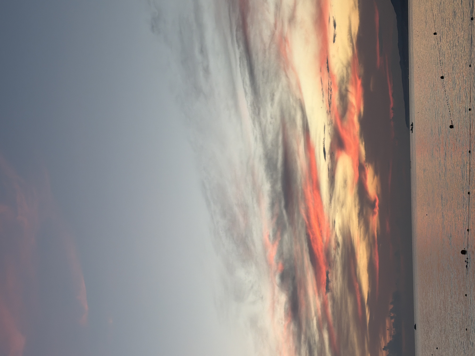 The image depicts a serene seascape at sunset over the Black Sea near Sochi, Russia. The sky is a canvas of soft gradients, transitioning from a pale blue at the top to vibrant hues of pink, orange, and yellow near the horizon, illuminated by scattered, wispy clouds. The sea below is calm and reflects the colors of the sky, creating a tranquil atmosphere. In the distance, a silhouetted mountain range lines the horizon. A small boat is visible on the water, adding a touch of life to the vast expanse. The foreground shows a roped-off area with buoys, suggesting a designated swimming or recreational zone. There are no people visible in the frame, emphasizing the peaceful and solitary nature of the scene.