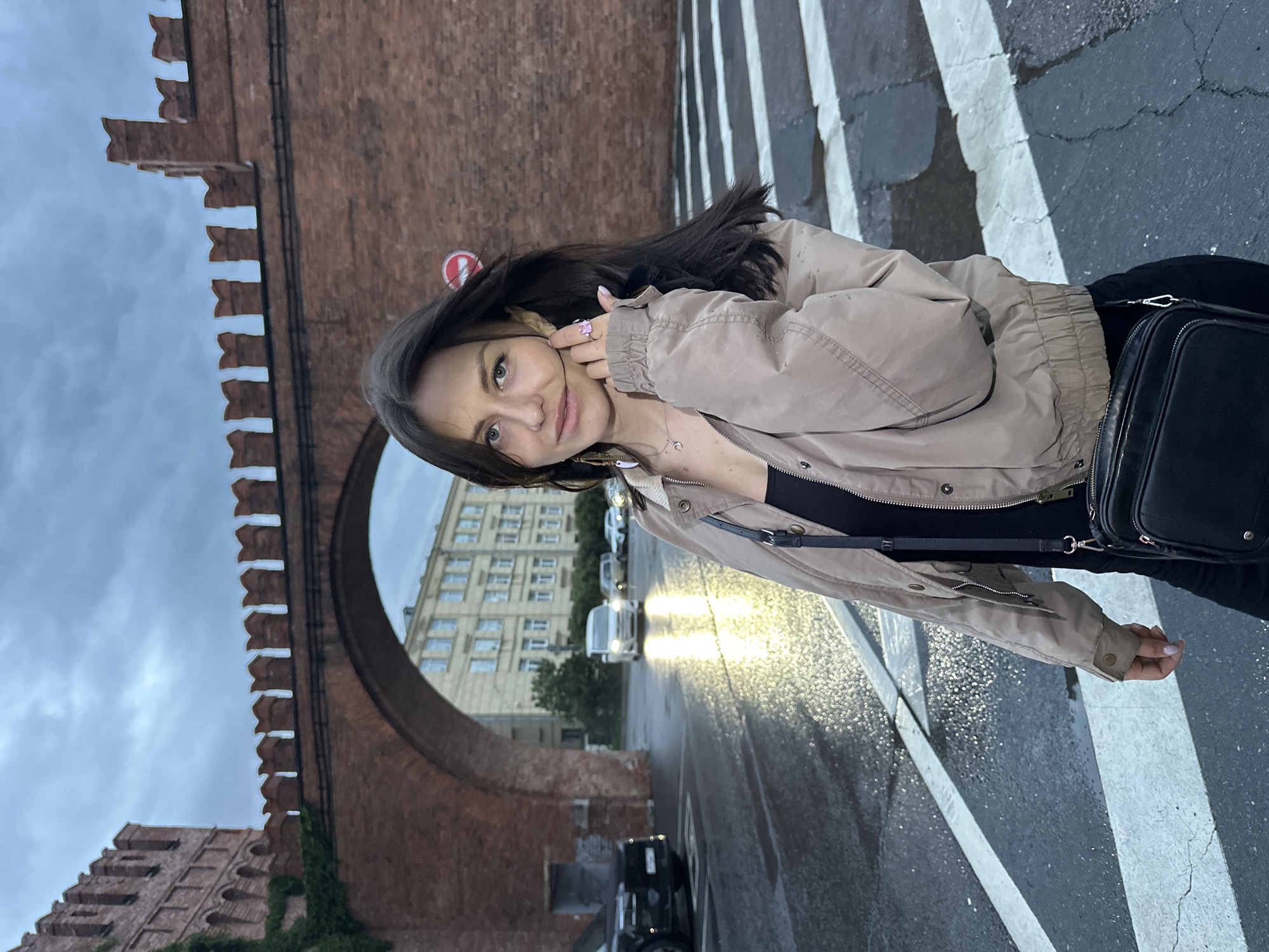 A young woman stands on a wet crosswalk in front of a large brick archway and building, likely in Saint Petersburg, Russia, given the architectural style. The archway appears to be part of a historic structure, possibly a gate or fortification, with crenellations along the top. The sky is overcast and cloudy. Behind the archway, a multi-story building with visible windows is illuminated, suggesting it is either daytime or the building has interior lights on. The street is wet, reflecting the headlights of vehicles. Two cars are visible on the left, and a white van or SUV is in the middle of the road. The woman is wearing a beige jacket over a black top and black pants. She has dark hair, and is wearing earrings, a necklace, and a ring. A black crossbody bag is slung over her shoulder. She is looking towards the camera and touching her face. The crosswalk lines are white and visible on the wet asphalt. A red and white circular traffic sign is partially visible to the right of the archway.