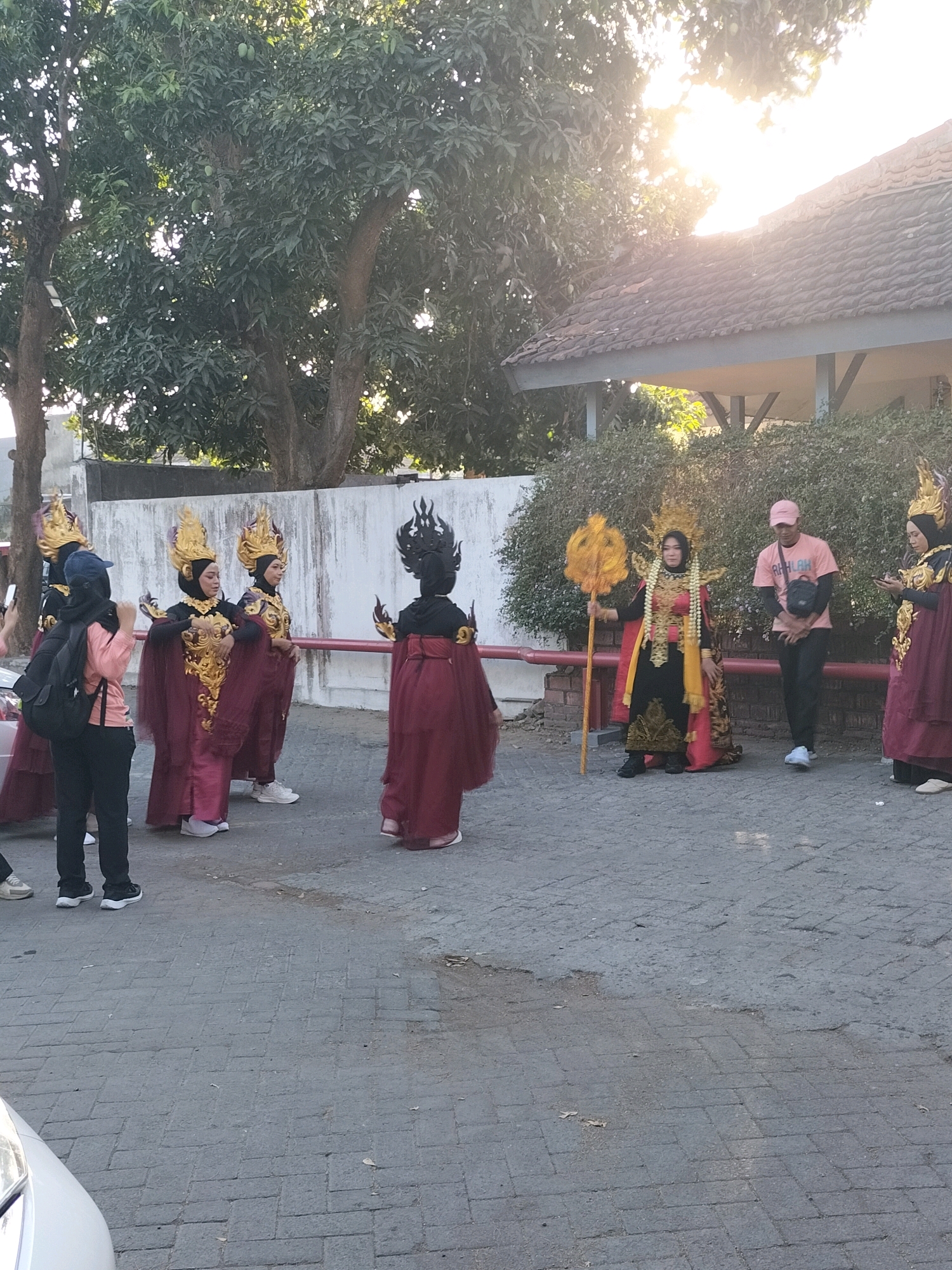 The image depicts an outdoor scene in Pare, Indonesia, likely a street or courtyard, with several individuals dressed in elaborate, traditional attire. In the foreground, a person in a pink shirt and a pink cap stands next to another person in traditional costume. The central group consists of individuals in striking, ornate outfits characterized by gold headdresses, richly colored robes in shades of maroon and gold, and intricate detailing. One individual in this central group holds a golden scepter or staff. To the left, two individuals are partially visible, also in traditional attire. The background shows a building with a tiled roof, dense foliage from trees, and a white wall. The lighting suggests it is daytime, with sunlight filtering through the trees, creating a bright, slightly overexposed effect in some areas. The ground is paved with interlocking bricks.
