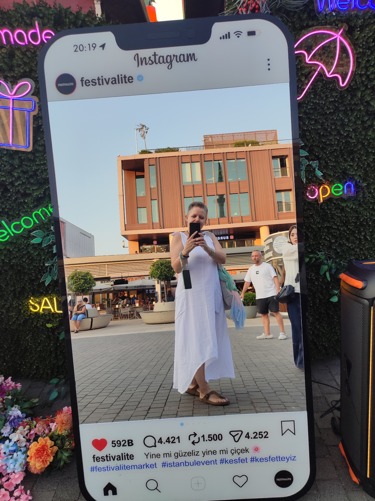 An outdoor scene in Istanbul, Turkey, possibly in the Beyoğlu district, is captured through a large, framed Instagram interface. The interface displays a selfie taken by a woman in a white dress, holding her phone. She stands on a paved area with a building with modern architecture in the background. To the left of the frame, neon signs read "made," "welcome," and "SALE." On the right, a neon sign displays an umbrella, and further down, another neon sign spells "open." The bottom of the Instagram interface shows likes, comments, and hashtags. A cluster of colorful flowers is visible in the lower left corner, and a portable speaker is on the right. The overall atmosphere suggests a lively public space, possibly a market or a commercial area, decorated with vibrant signage and artificial greenery.