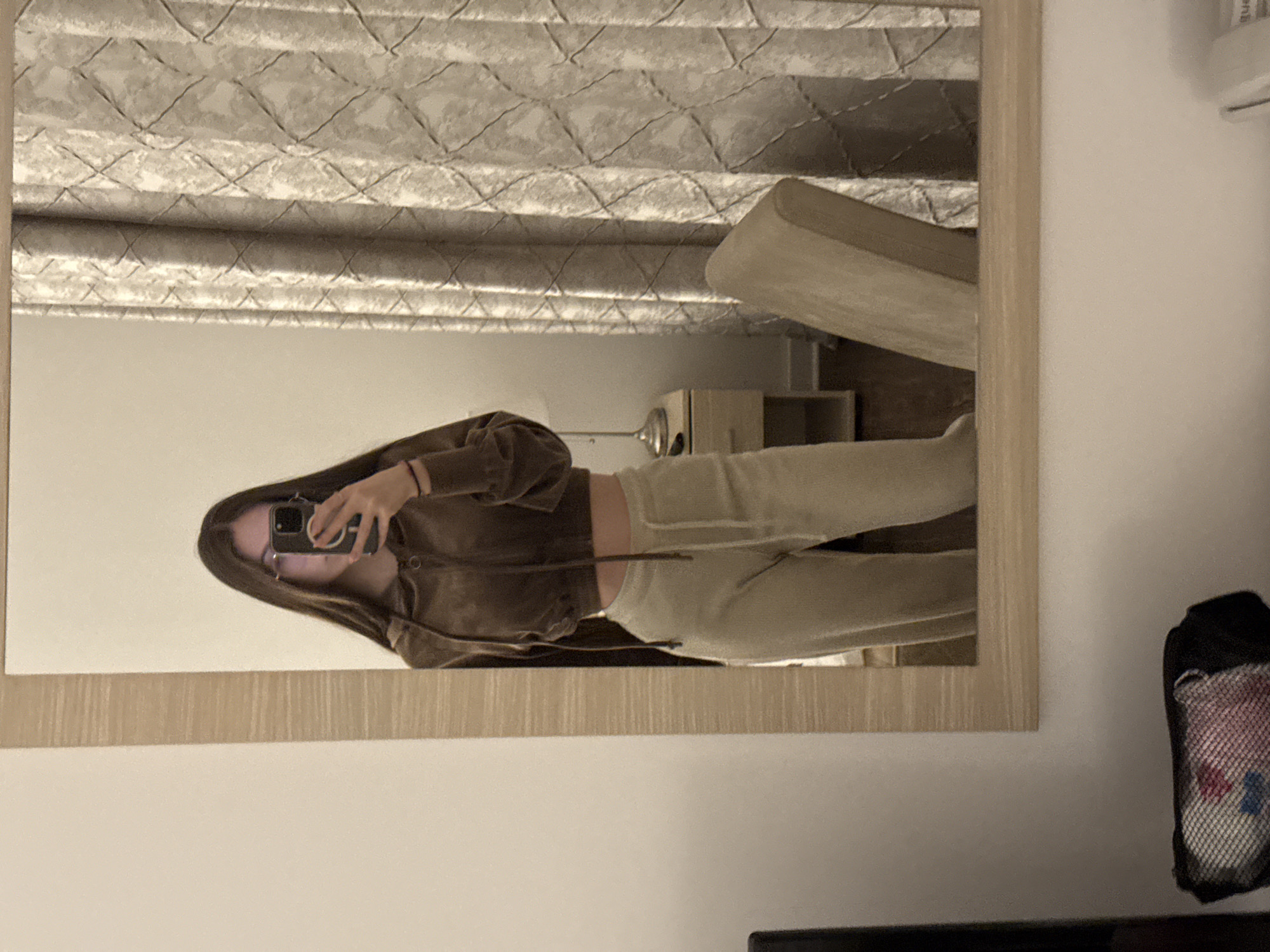 The image depicts a female subject capturing her reflection in a rectangular mirror with a light wood-toned frame, presumed to be located within the city of Vozdovac Urban Municipality, Serbia. The subject, possessing long dark hair and wearing glasses, is partially obscured by a dark-colored smartphone, held to photograph her upper body. She is dressed in a brown long-sleeved cropped hooded sweatshirt and light beige sweatpants. A black bracelet or band is visible on her left wrist.
The reflection in the mirror reveals a room interior. To the subject's left is a plain white wall. To her right, a light-colored curtain with a repetitive geometric (crisscross/diamond) pattern hangs. A small wooden bedside table with a silver-toned lamp is visible between the subject and the curtain, slightly to her left. A light-colored upholstered chair is positioned to her right, partially obscured by the mirror frame. In the lower left foreground, outside the mirror's reflection, a black mesh bag containing colorful items (red, blue, white) is present. The room is illuminated by warm, indirect lighting.