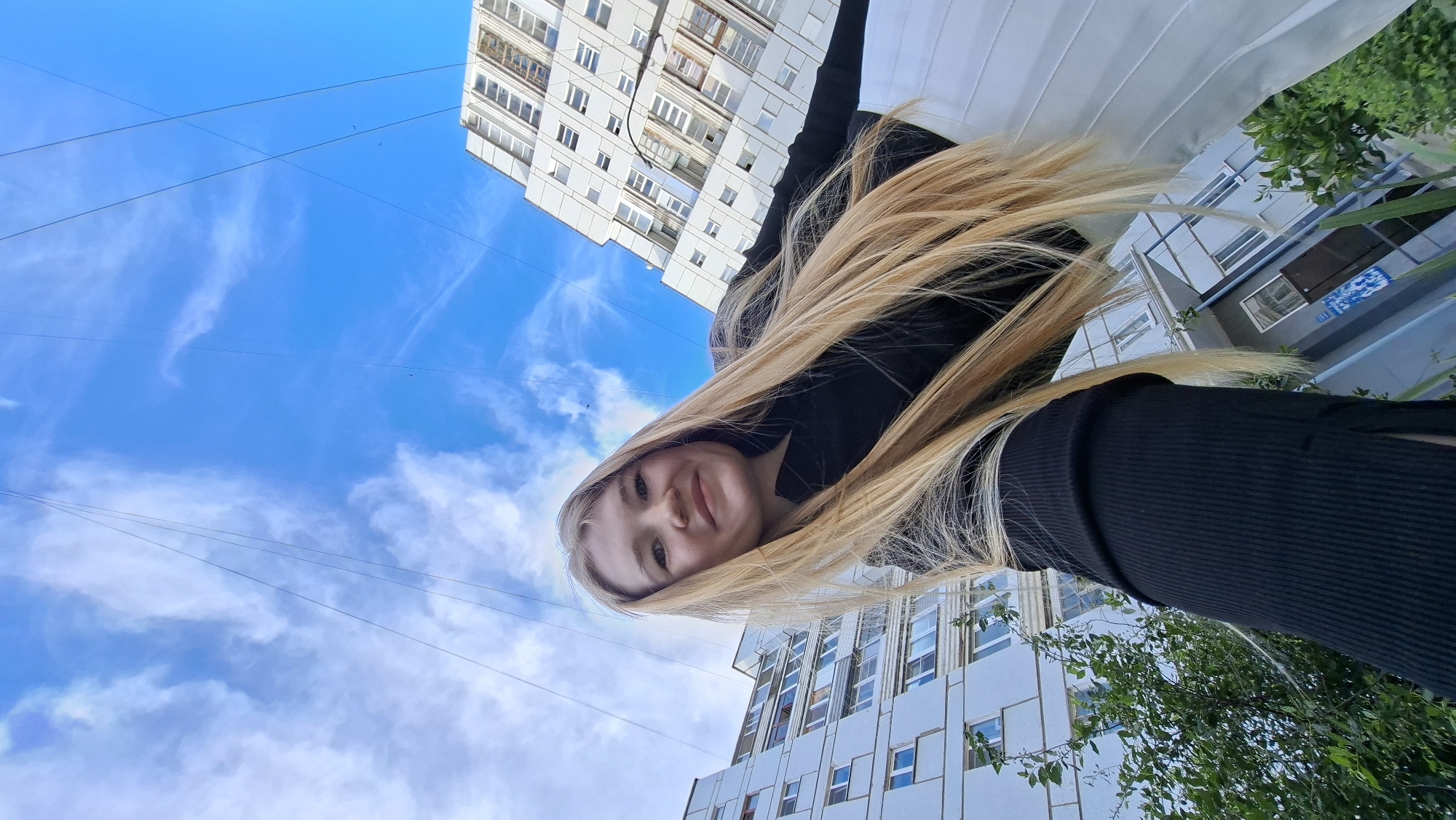 In a low-angle, wide shot taken on a cloudy day in Chaikovsky, Russia, a young woman with long, blonde hair smiles directly at the camera. She is wearing a black, ribbed, long-sleeved top and a white pleated skirt. Behind her, two large, light-colored apartment buildings loom under a bright blue sky with wispy white clouds. Several thin wires crisscross the sky above the buildings. Green foliage is visible at the bottom left and right of the frame, near the base of the buildings.