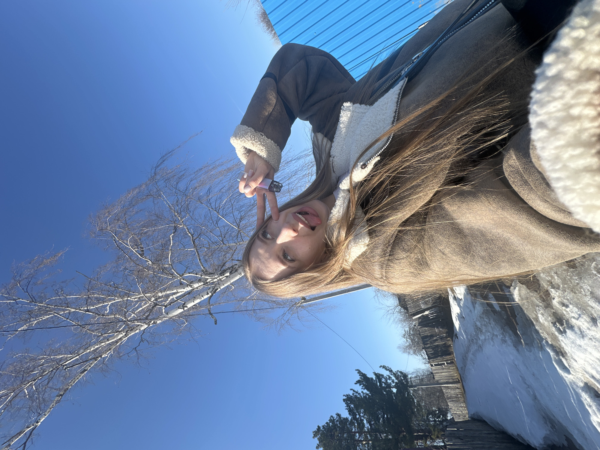A young woman with long blonde hair and blue eyes is pictured making a peace sign with her right hand while sticking out her tongue. She is wearing a brown suede jacket with a white shearling collar and lining. She is holding a small, pink disposable vape pen in her right hand.
The background is a bright blue sky with a bare birch tree on the left. In the bottom right corner, there is a snow-covered wall with a wooden fence. The location is in the city of Chaikovsky, Russia.