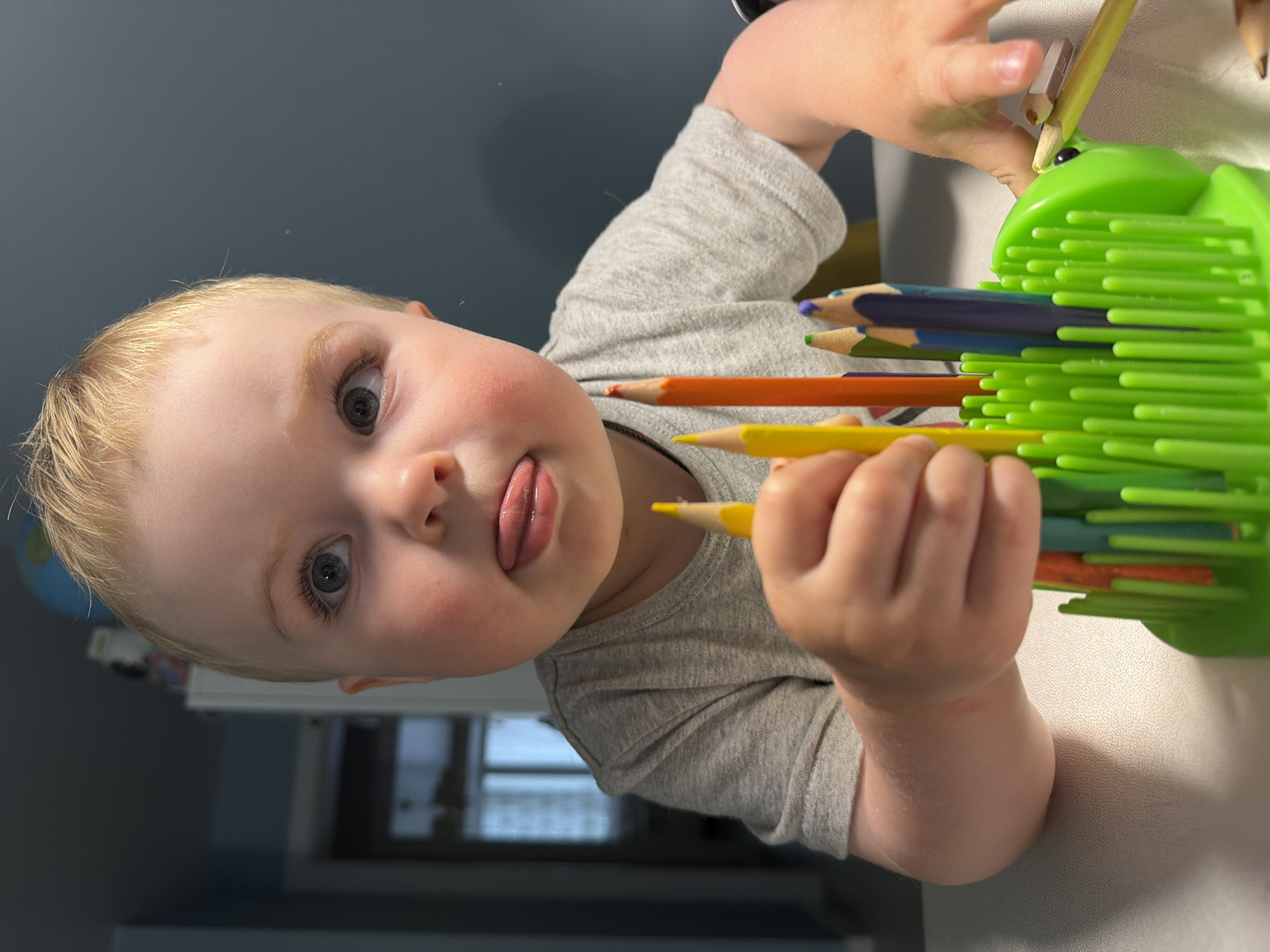 A young child with blonde hair and blue eyes is sticking their tongue out while holding an assortment of colored pencils. The pencils are partially inserted into a green holder with many flexible green extensions. The child is wearing a gray short-sleeved shirt. The location is unknown and there is no evidence to suggest that this photo was taken in Votkinsk, Russia.