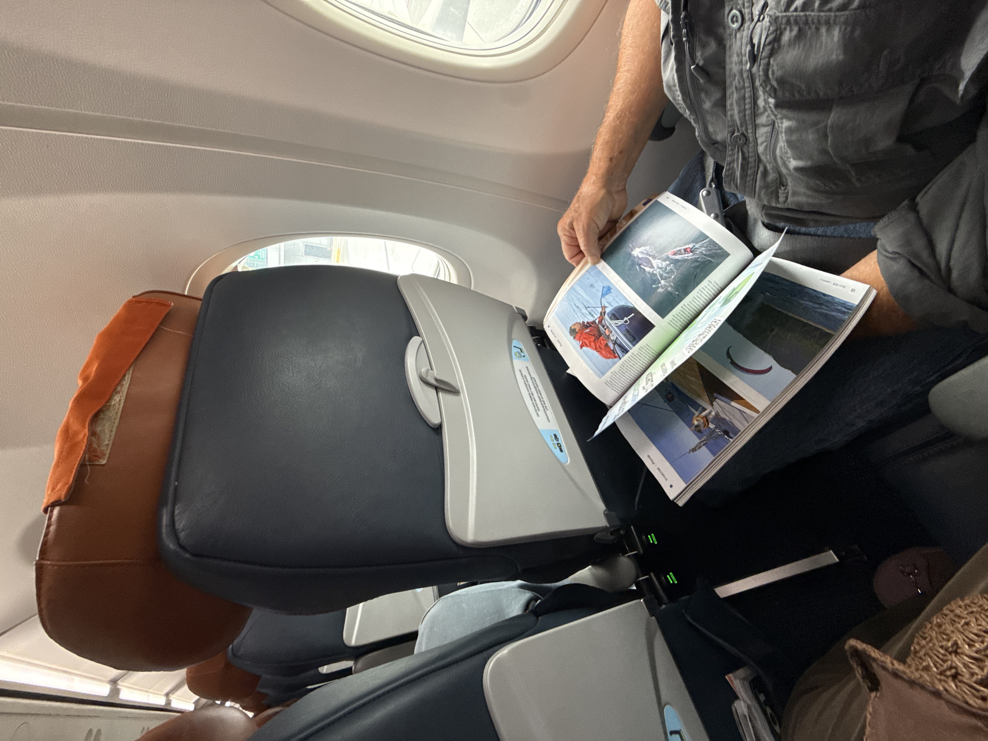 An elderly Caucasian male is seated in an airplane cabin, reading a magazine. He is wearing a grey t-shirt and grey shorts. The magazine is open to pages featuring images of people engaged in water sports. The man's hands are visible, holding the magazine. The seats in front of him are dark grey with light grey headrests. To the left, another airplane seat is visible. The aisle between the seats has charging ports. The image is taken from a slightly elevated perspective, looking down towards the man and his magazine. The environment suggests an indoor setting, likely an airplane on the ground or in flight, given the presence of airplane seats and the view out the window which shows what appears to be an airport tarmac or terminal building. The lighting is neutral.