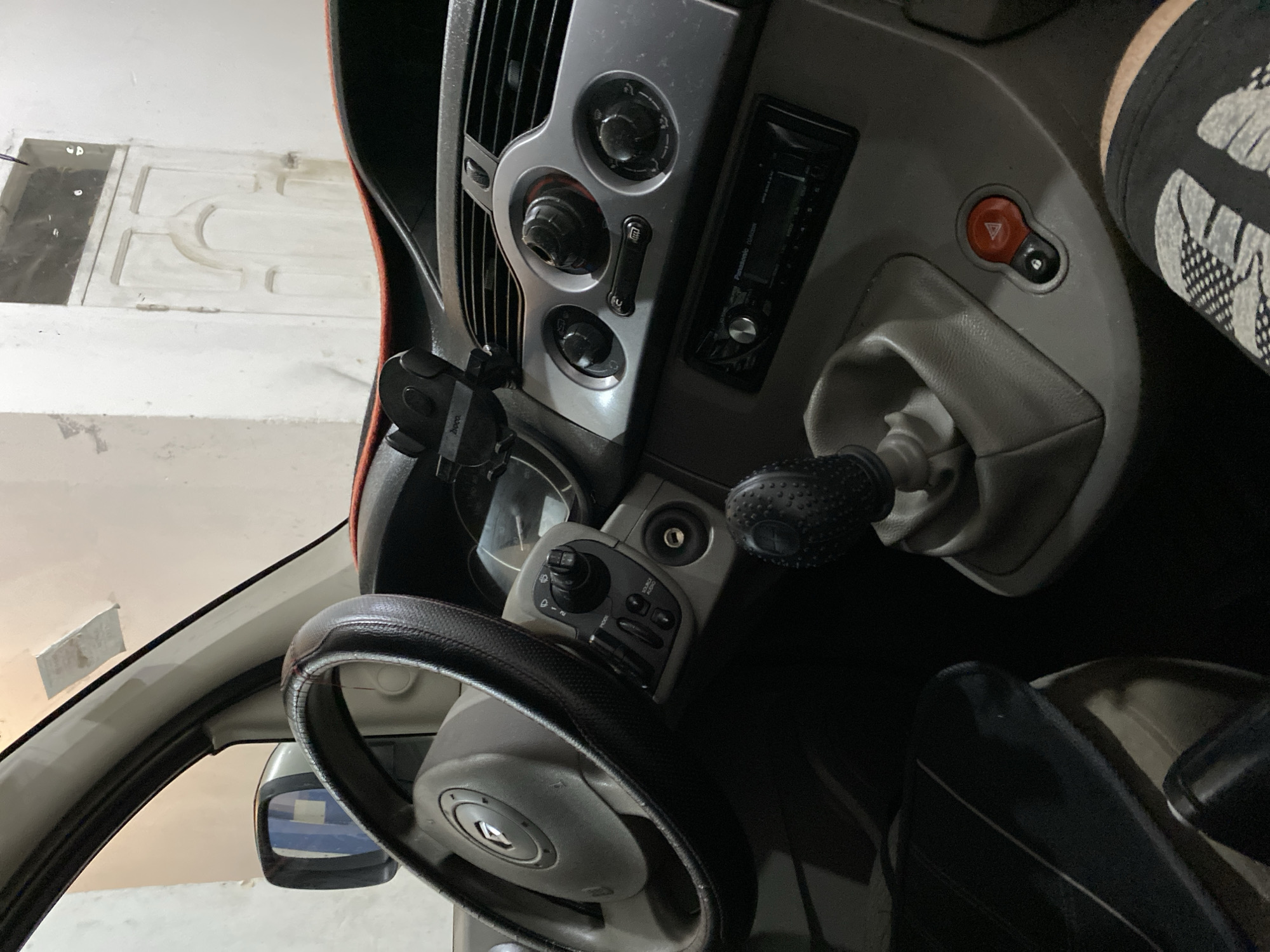 The image shows the interior of a vehicle, specifically the dashboard, steering wheel, and gear shift. The dashboard is a light gray color with silver accents. The steering wheel has a black cover. The gear shift knob is also black and textured. Part of the driver's leg is visible, wearing black patterned athletic wear. The environment appears to be an indoor parking structure or garage, as suggested by the concrete wall and door in the background. The location in Baraki, Algeria is not directly verifiable from the image alone, as there are no distinctive visual indicators to confirm this.