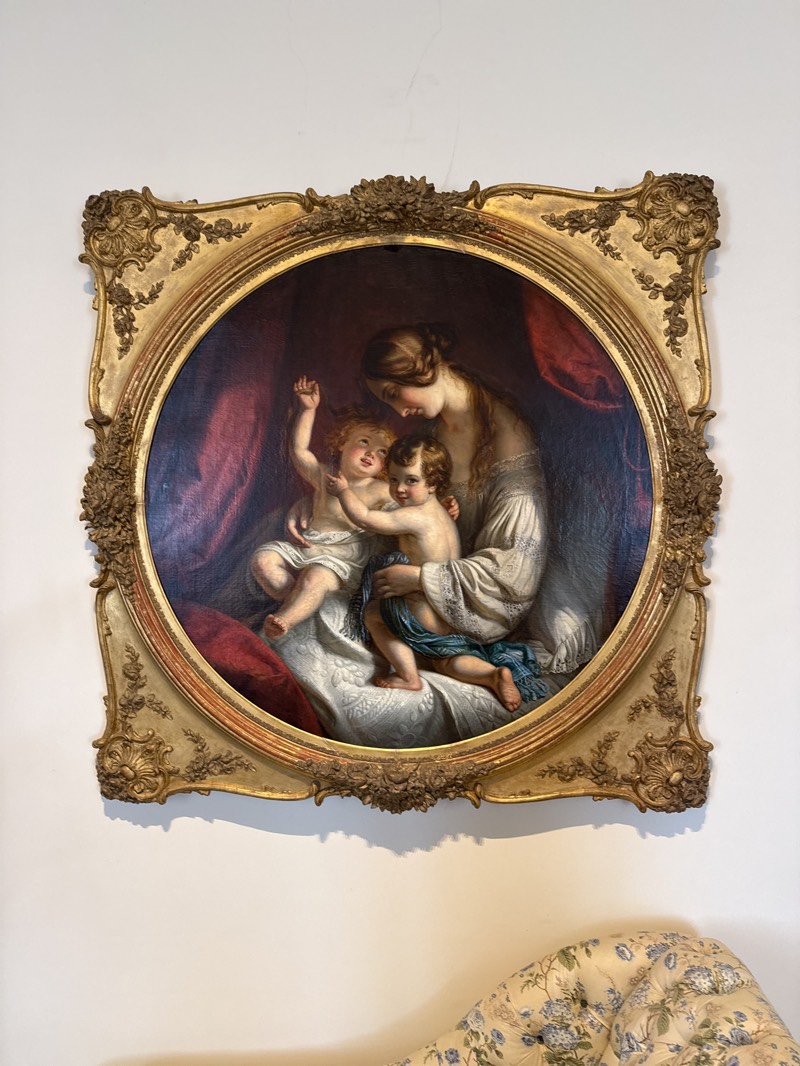 The image depicts a framed painting hanging on a white wall. The painting is round, framed in a detailed, ornate gold frame. The painting itself shows a woman in a light-colored dress holding two cherubic children. One child is reaching up with an outstretched arm, while the other nestles into the woman's arms. A red drape provides a backdrop to the figures. The bottom portion of the image reveals the top of a floral-patterned chaise lounge. It is probably located at the Pena Palace, a romanticist castle in São Pedro de Penaferrim, in the municipality of Sintra, Portugal.