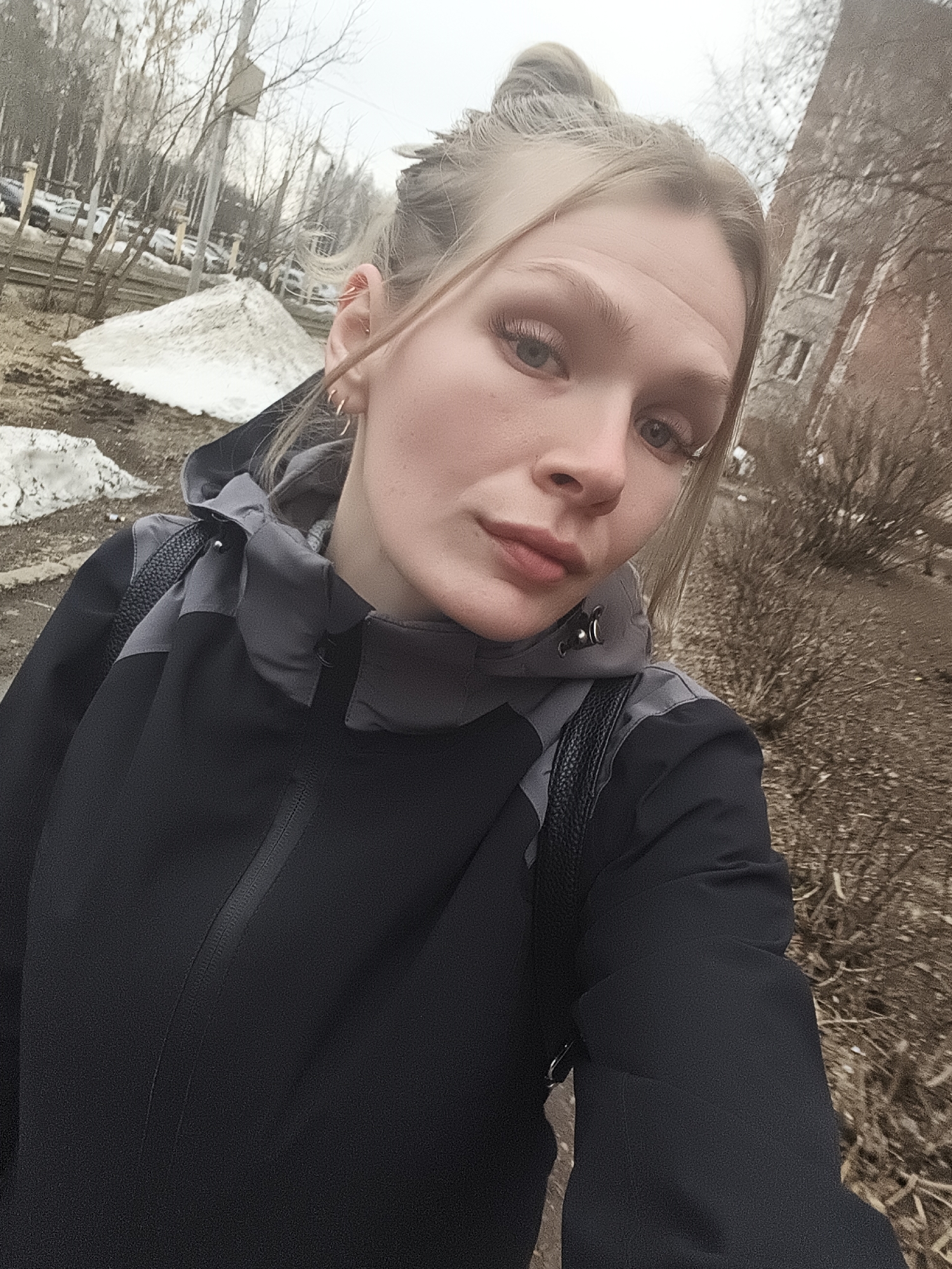 A young woman with blonde hair pulled back in a bun is taking a selfie outdoors. She is wearing a black jacket with a gray hood and has a black leather backpack. The background shows signs of early spring; patches of melting snow, bare trees, and brown foliage are visible. The location appears to be Izhevsk, Russia, given the architecture and typical urban landscape. A multi-story brick building can be seen on the right side, and several parked cars are visible in the background.