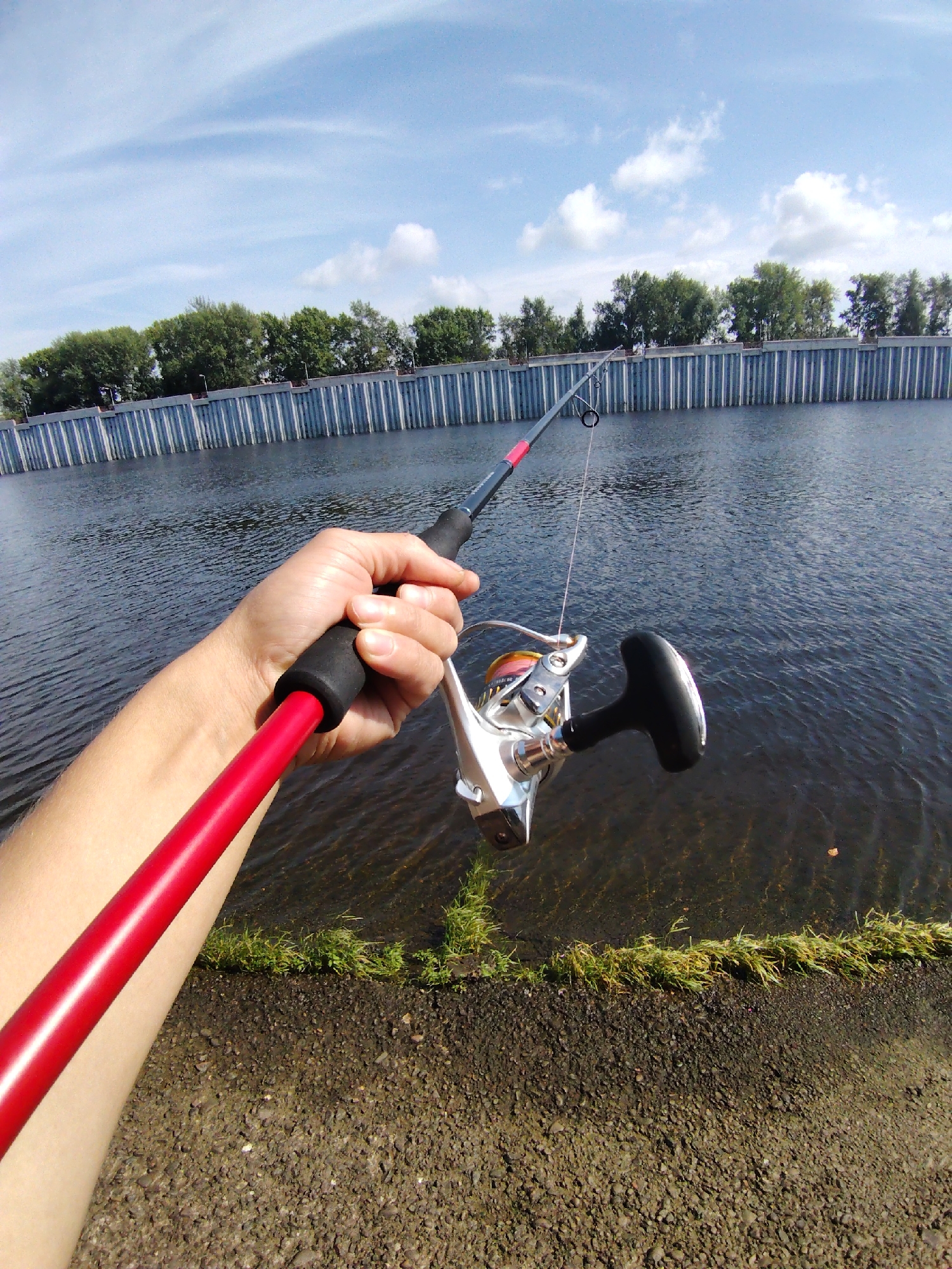 A first-person perspective shot shows a hand holding a fishing rod over the water. The rod is red and black with a silver reel. The background features a concrete wall with trees behind it, and a blue sky with scattered clouds. The foreground shows the ground, which is a mix of gravel and grass. The scene appears to be a waterfront area in Chaikovsky, Russia, possibly near the Kama River.